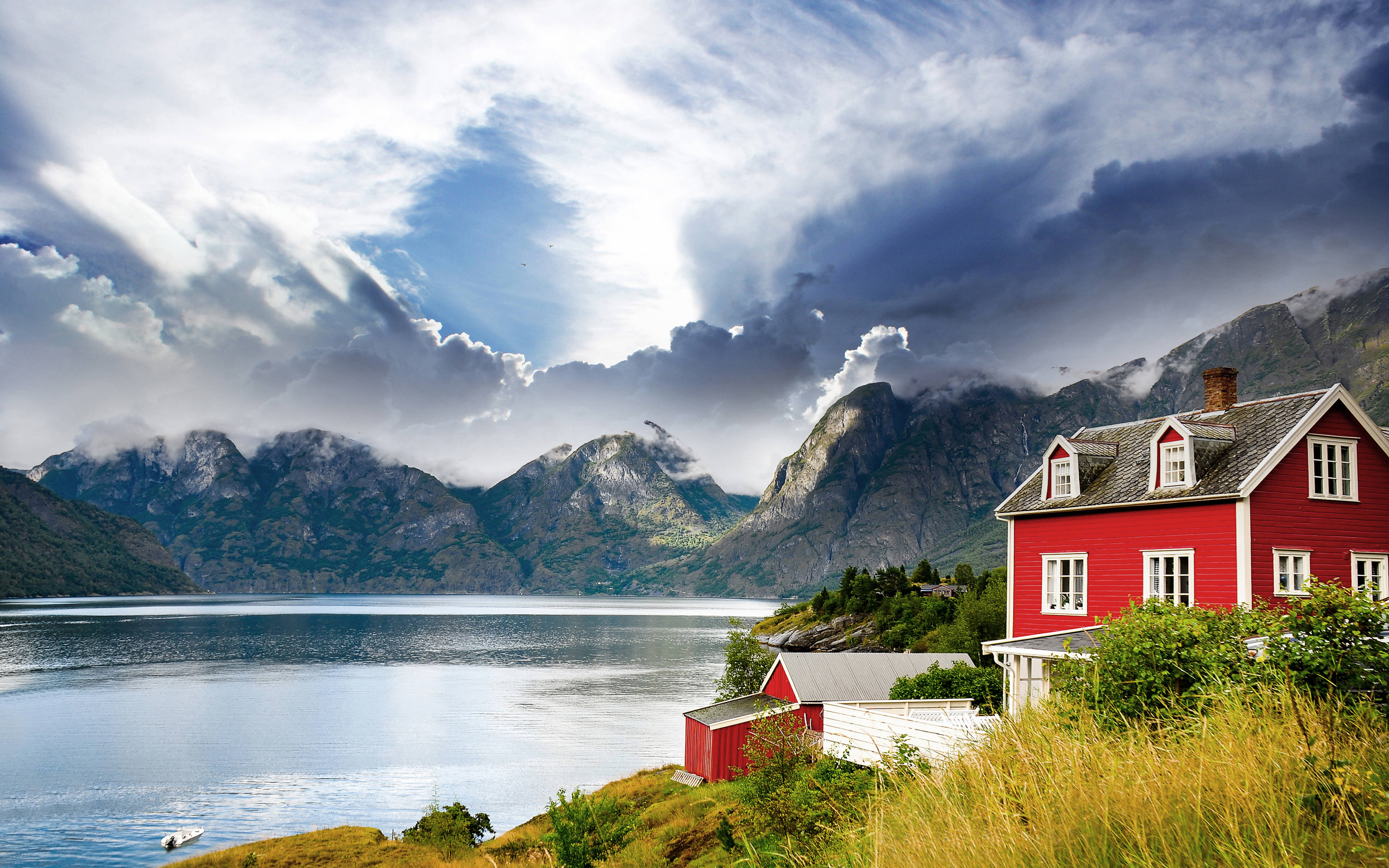 Norway 桌布 壁紙 — 一棟鮮紅色的房子坐落在長滿青草的懸崖上，俯瞰著寧靜的峽灣，霧氣繚繞的山脈在戲劇性的多雲天空下。