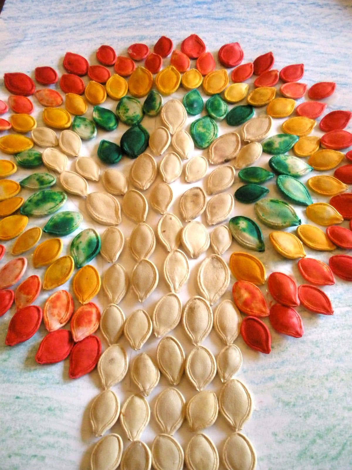Fall Crafts 桌布 壁紙 — 一棵充滿活力的樹，由塗色的南瓜籽排列成彩虹色，放在有紋理的白色表面上。