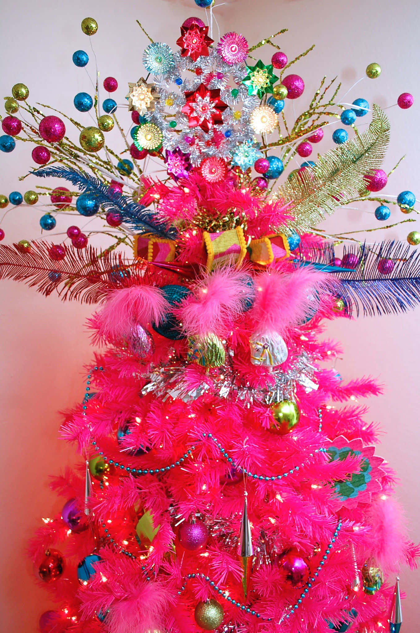 Pink Christmas Tree 桌布 壁紙 — 一棵充滿活力的粉紅色人造聖誕樹，飾有閃亮裝飾品、羽毛和彩色飾球，襯著柔和的粉紅色牆壁。