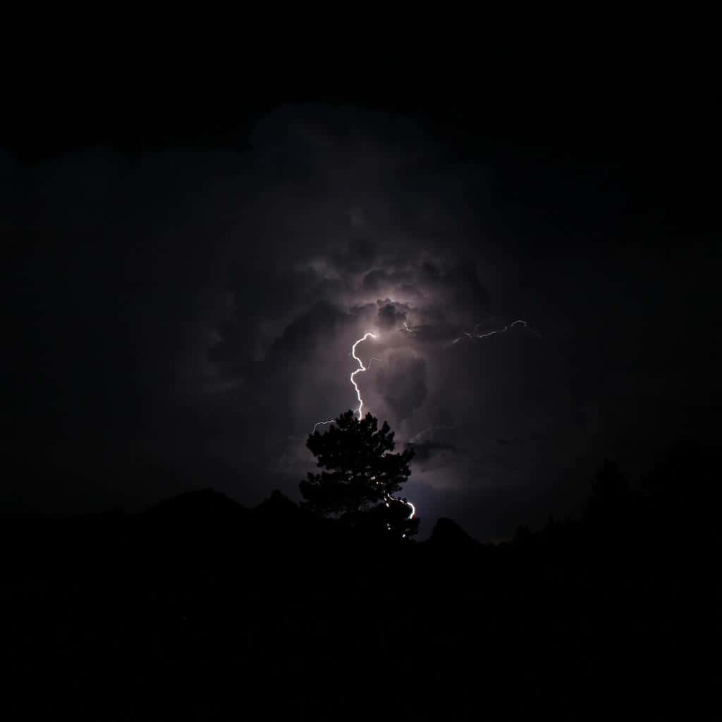 Thunderstorm 桌布 壁紙 — 一棵孤獨的松樹在黑暗的天空中形成剪影，一道明亮的鋸齒狀閃電擊中其頂部附近。
