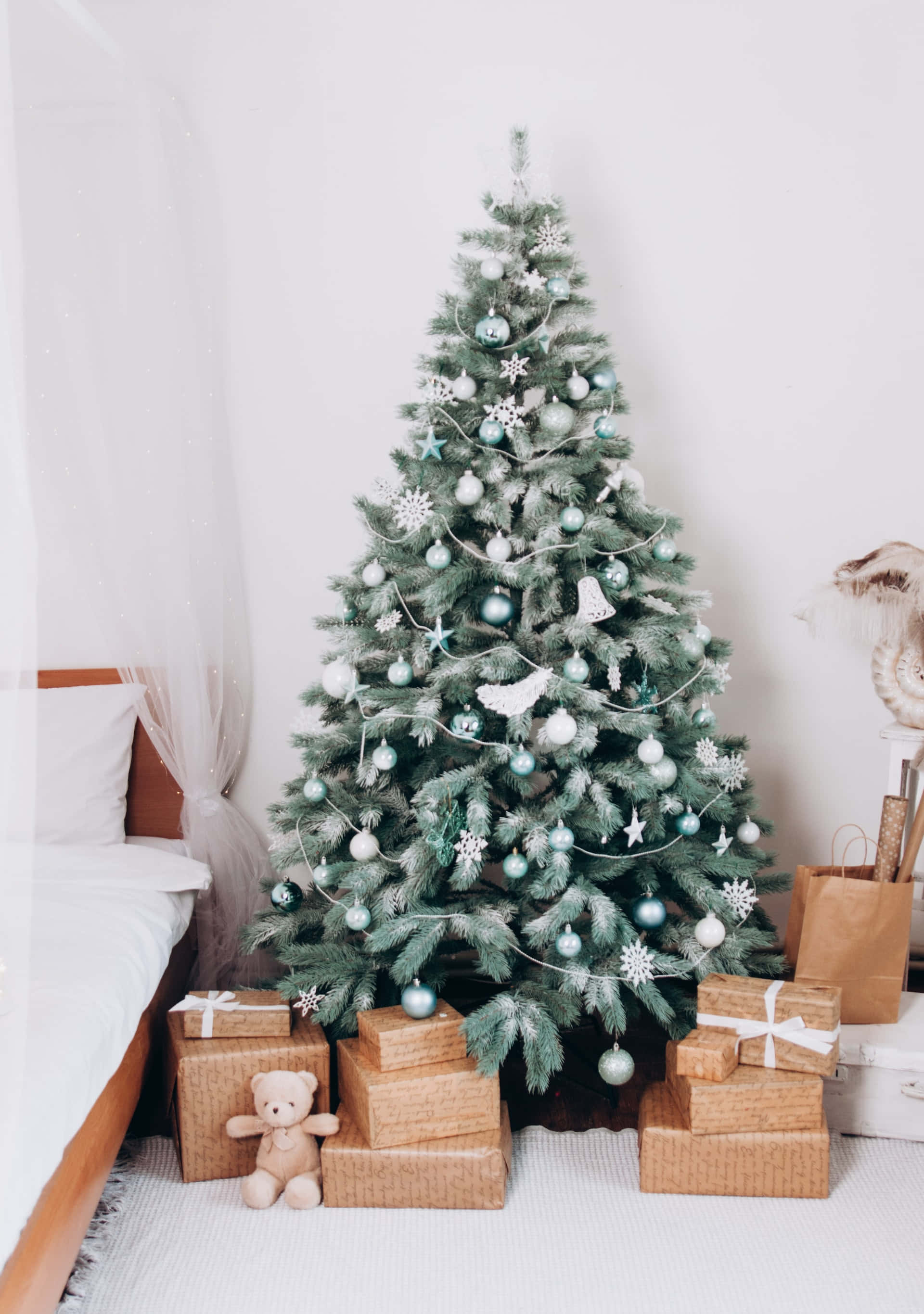 Aesthetic Christmas Tree 桌布 壁紙 — 一棵植絨聖誕樹裝飾著青綠色和白色的飾物，旁邊有一張床，床腳放著包裝好的禮物和一隻泰迪熊。