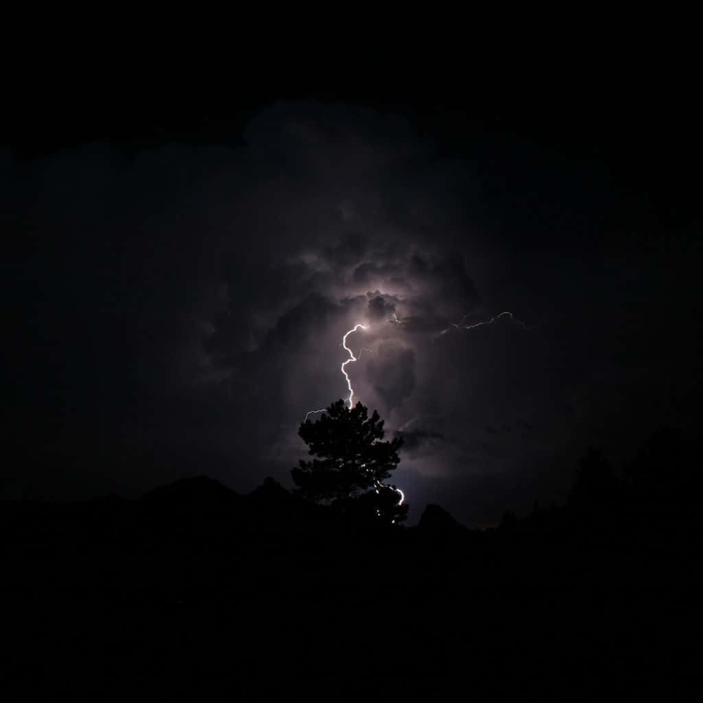 Black And White Storm 桌布 壁紙 — 一棵樹在黑暗天空下的剪影，一道明亮的閃電擊中其頂部附近。