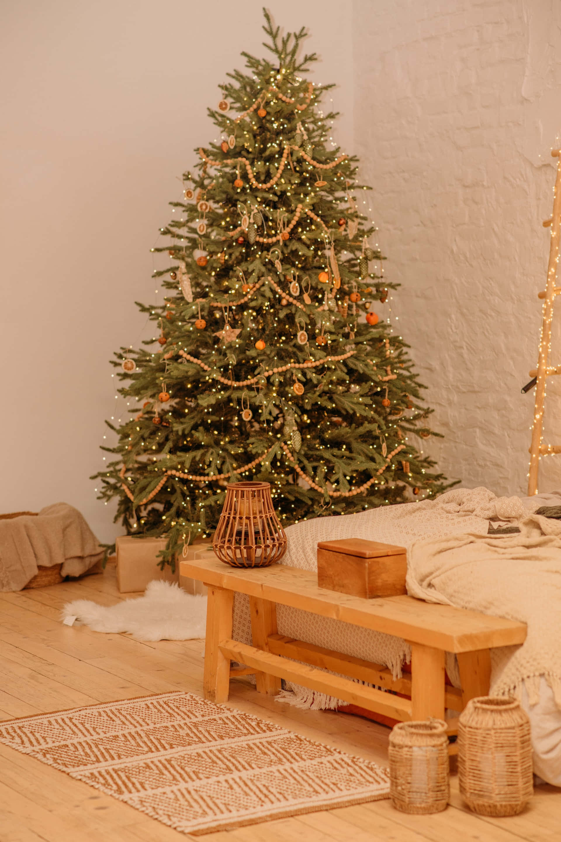 Aesthetic Christmas Tree 桌布 壁紙 — 一棵溫暖照亮的聖誕樹，裝飾著燈串和木製飾品，旁邊放著一張木長椅，位於舒適的鄉村風格房間內。