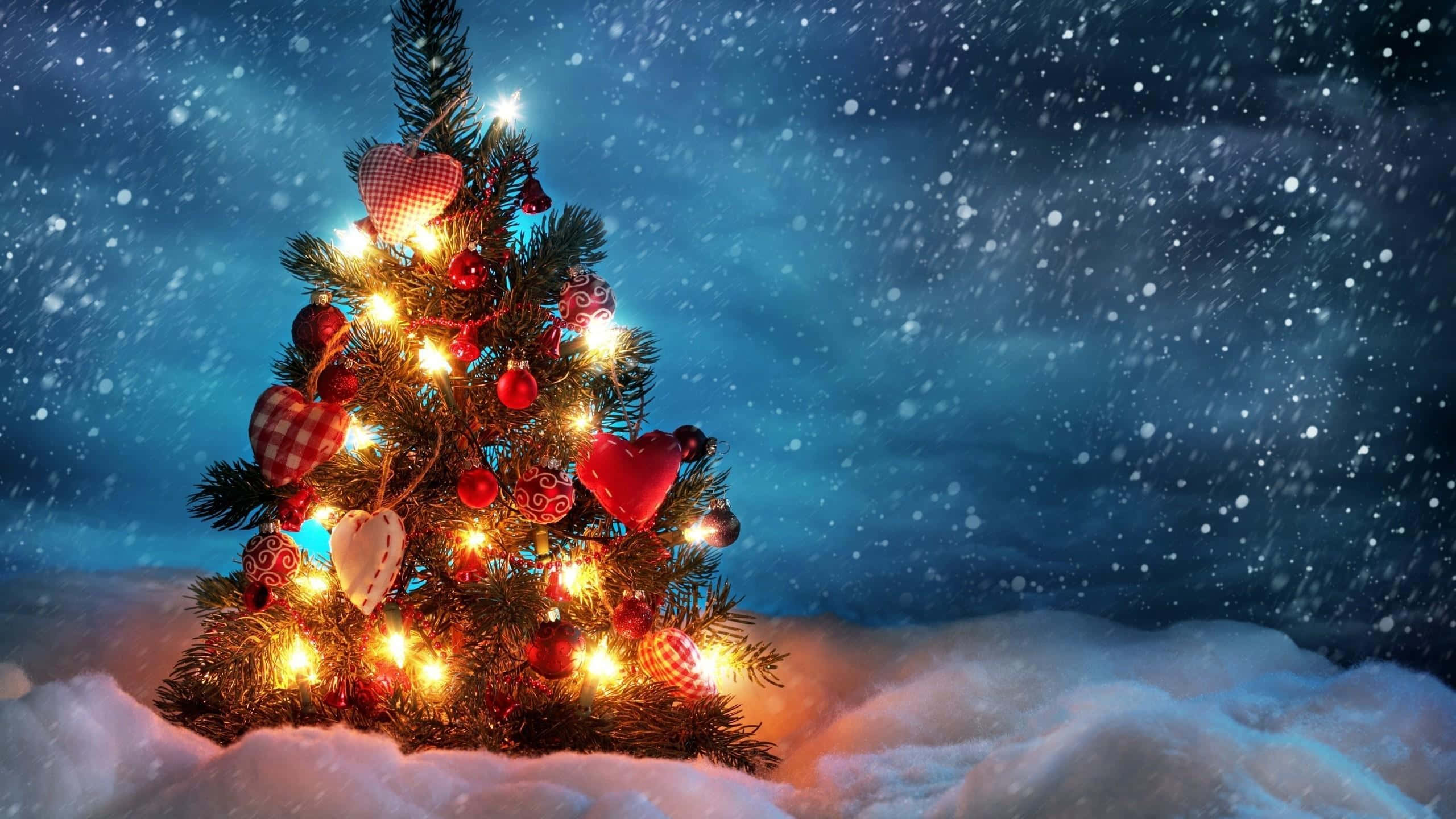 1920x1080 HD Christmas 桌布 壁紙 — 一棵溫暖照亮的聖誕樹，裝飾著紅色心形和飾物，矗立在雪地中，背景是繁星點點的夜空，雪花飄落。