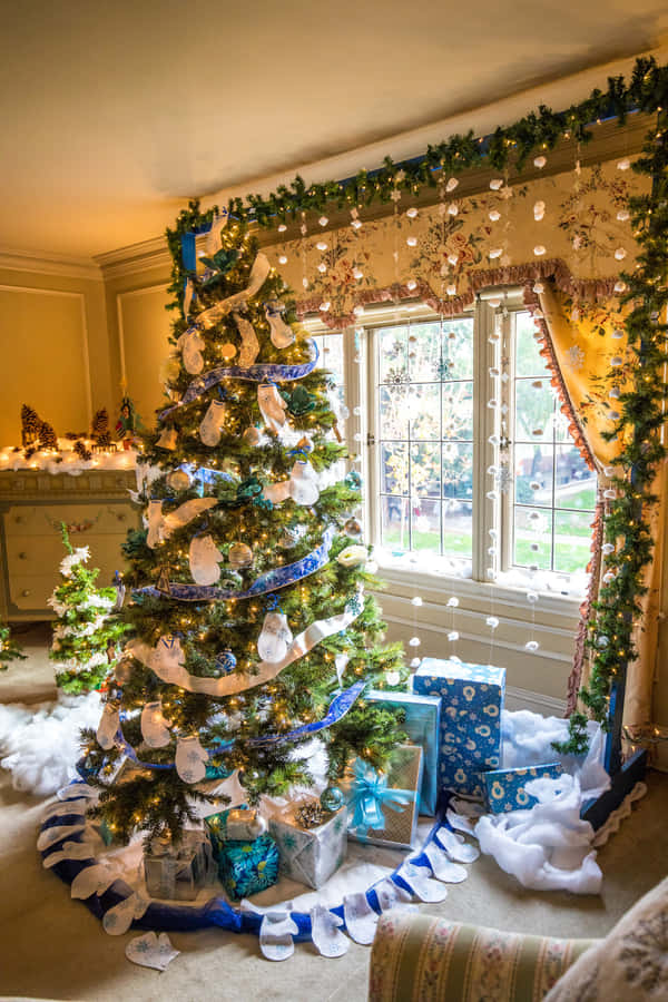 Aesthetic Christmas Tree 桌布 壁紙 — 一棵溫暖照亮的聖誕樹，裝飾著藍色絲帶、白色襪子和禮物，放在窗下，窗戶掛滿節日花環和仙女燈。