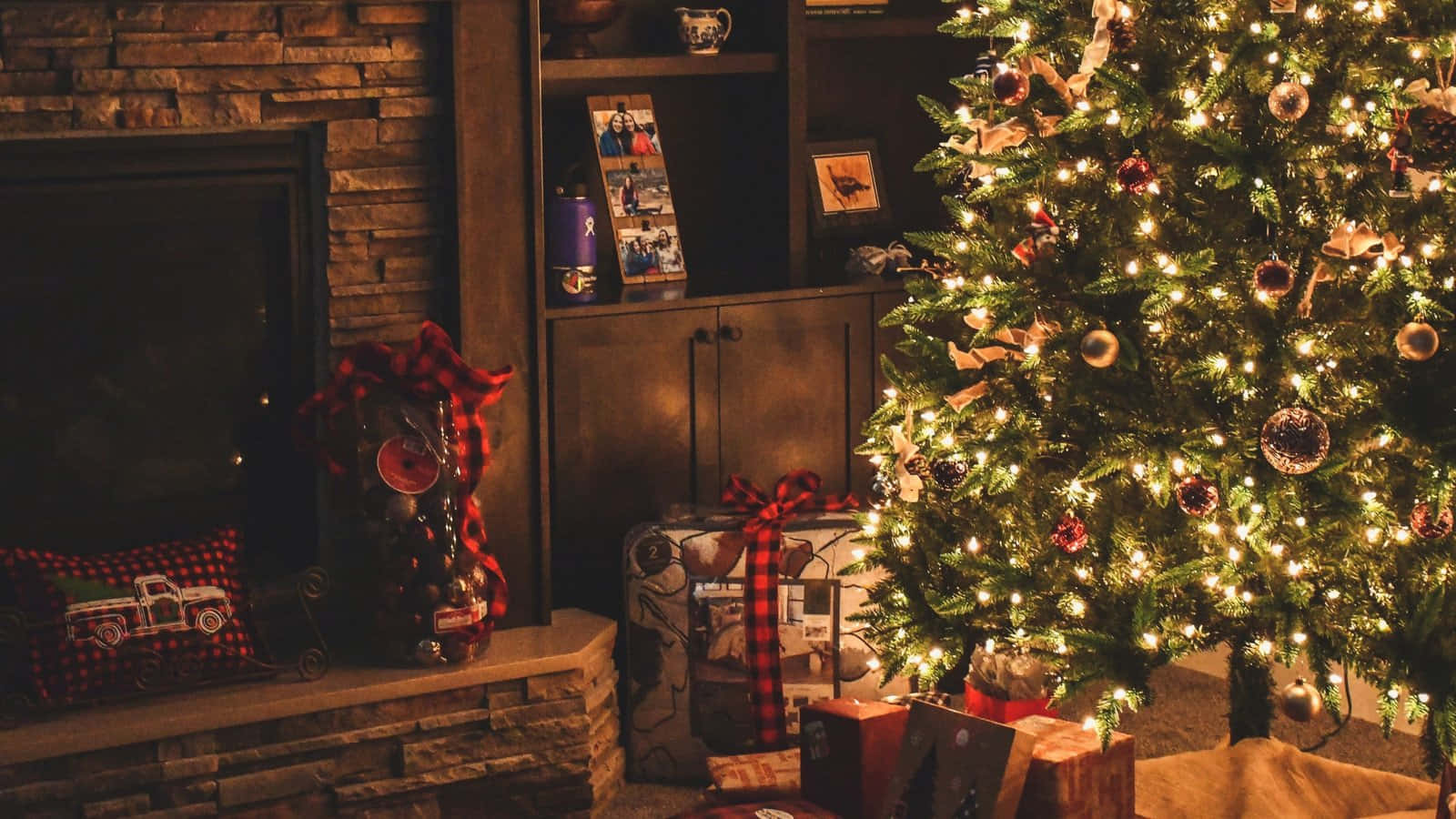 Teams Christmas 桌布 壁紙 — 一棵溫暖照亮的聖誕樹，裝飾著飾品和燈光，站在石壁爐旁，旁邊有節日禮物和格子裝飾。