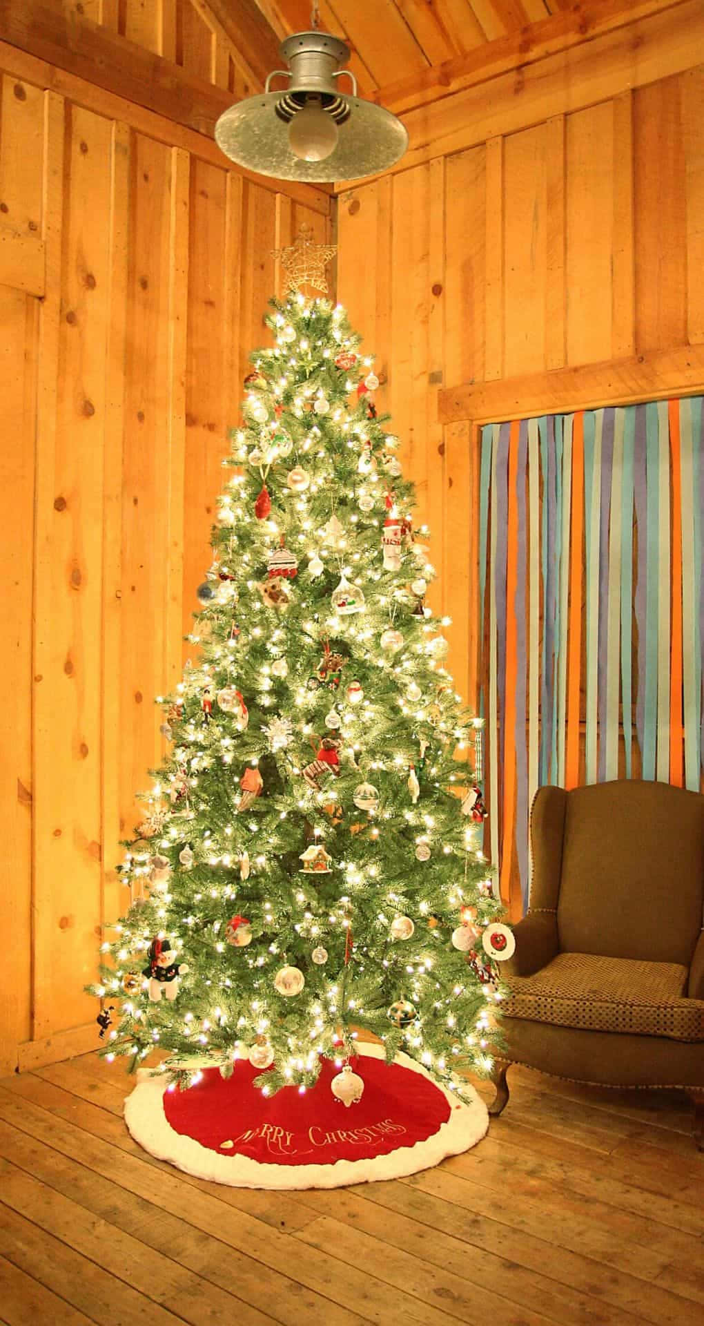 Aesthetic Christmas Tree 桌布 壁紙 — 一棵溫暖照亮的聖誕樹，裝飾著飾品和燈光，矗立在舒適的木屋角落，旁邊有一張椅子。