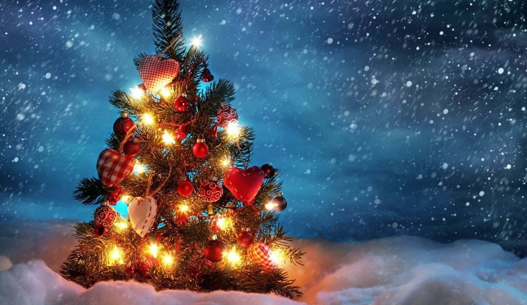 Aesthetic Christmas Tree 桌布 壁紙 — 一棵溫暖照明的聖誕樹，裝飾著紅色心形和飾品，矗立在雪地中，背景是雪花飄落的夜空。