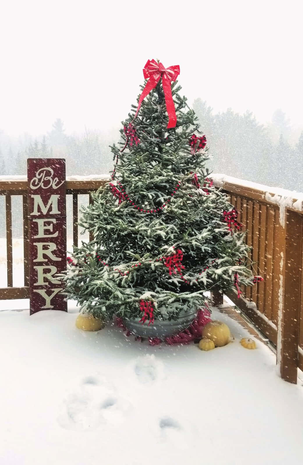  壁紙 — 一棵灑滿雪的聖誕樹，裝飾著紅色蝴蝶結和漿果，立在舖雪的甲板上，旁邊有一個「Be MERRY」標牌。