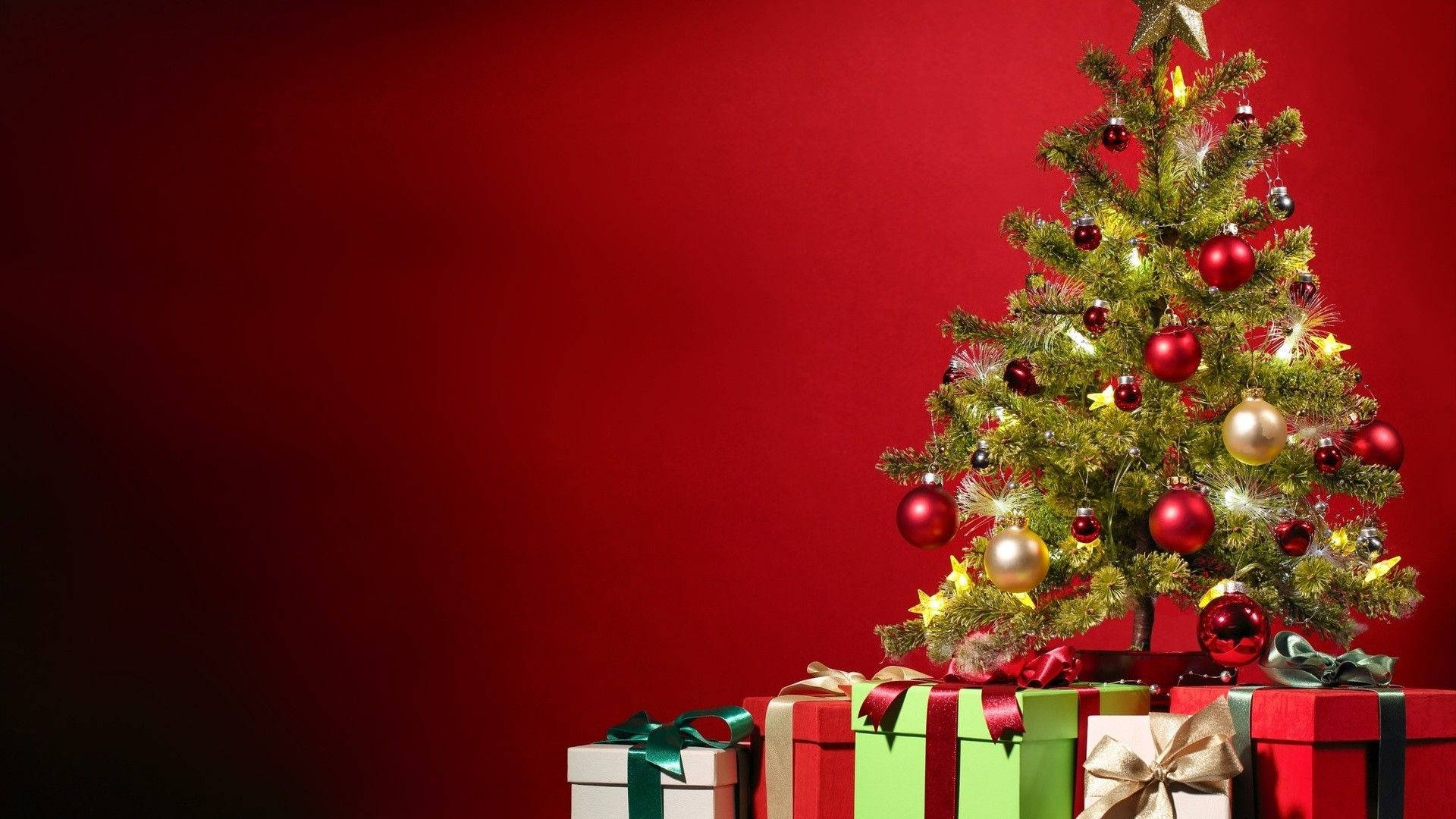 聖誕節 桌布 壁紙 — 一棵節日聖誕樹裝飾著紅色、金色和銀色的飾物，旁邊放著色彩繽紛的包裝禮物，背景是濃郁的紅色。