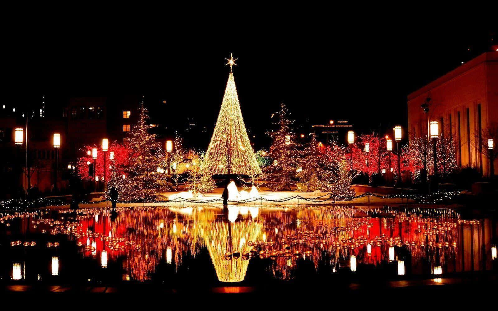 Christmas Eve 桌布 壁紙 — 一棵耀眼的聖誕樹在夜晚閃耀著金色的燈光，倒映在寧靜的池塘中，周圍是紅燈點綴的樹木和建築物。