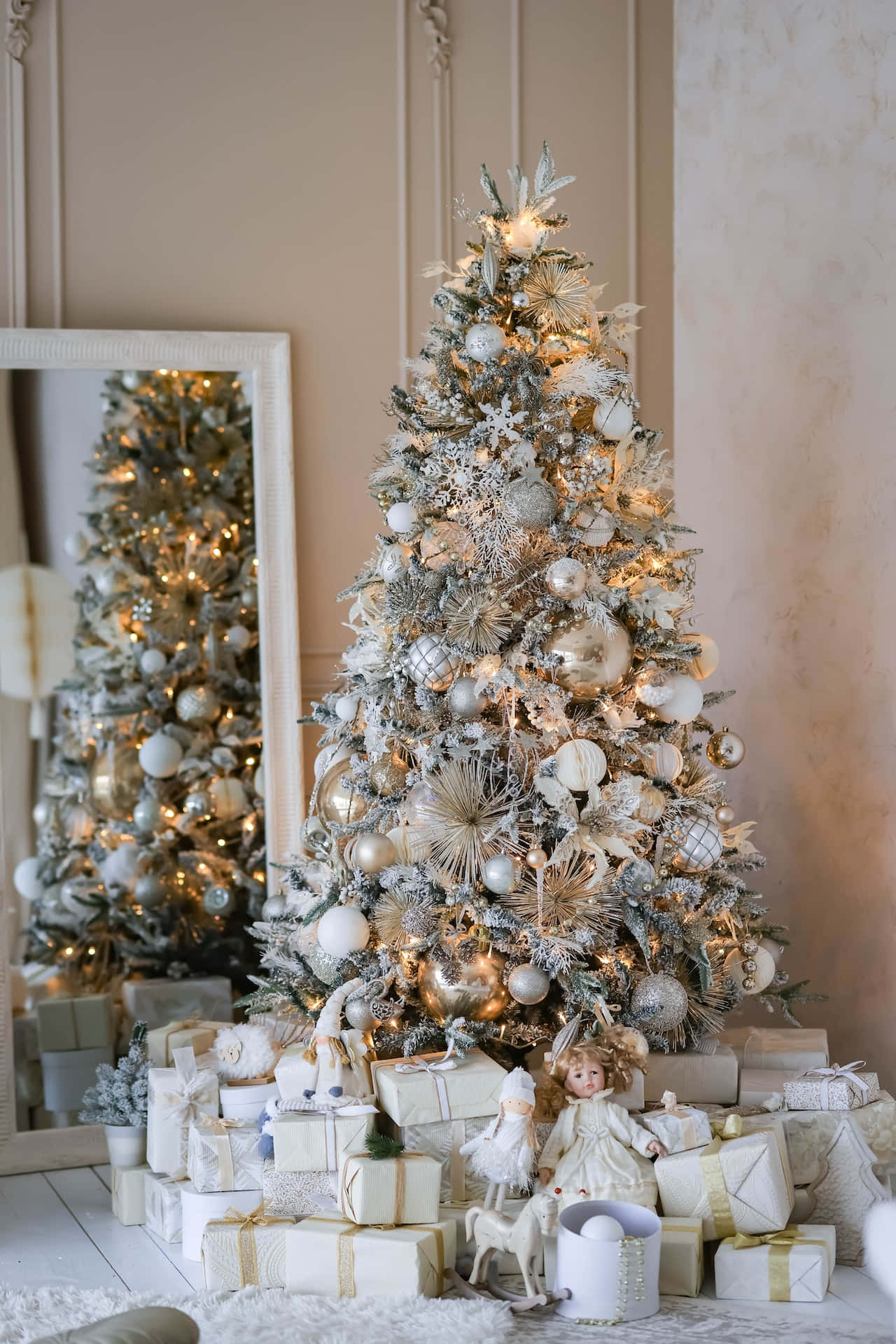 Aesthetic Christmas Tree 桌布 壁紙 — 一棵華麗裝飾的銀色和金色聖誕樹溫暖地閃爍著燈光，周圍環繞著包裝好的禮物和一個洋娃娃，營造出舒適的室內氛圍。