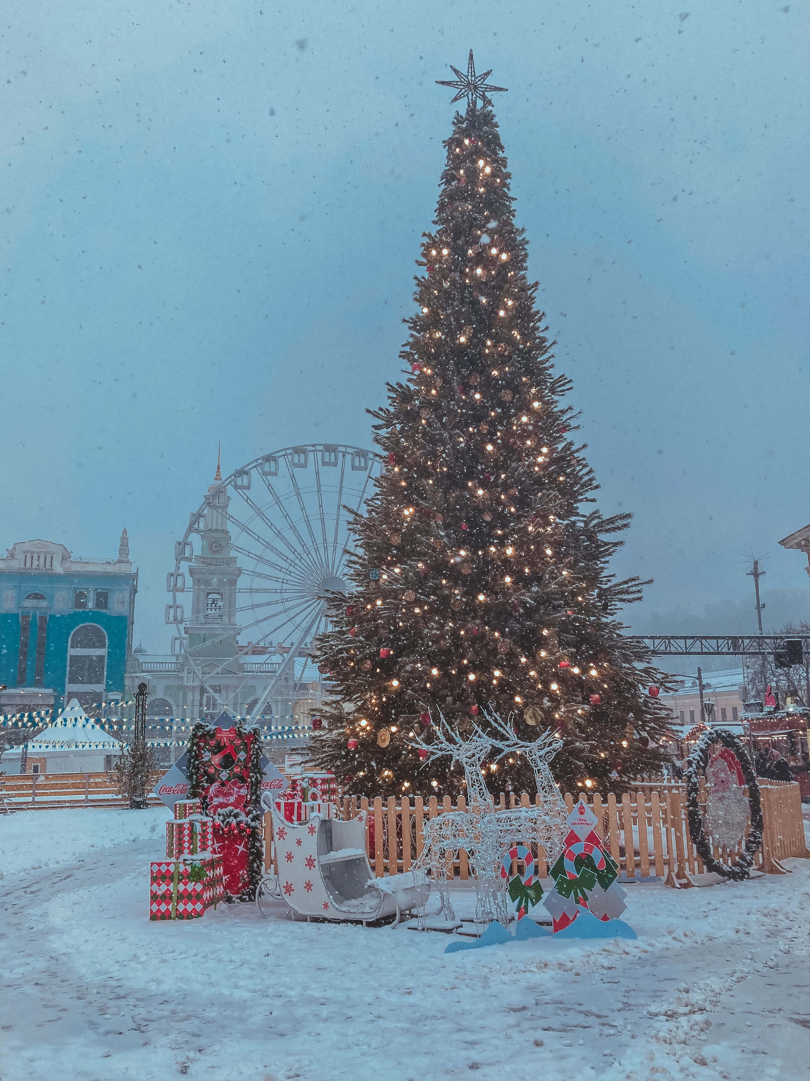 Christmas Scene 桌布 壁紙 — 一棵被雪覆蓋的聖誕樹，裝飾著燈飾和飾品，矗立在冬日廣場上，背景是摩天輪。