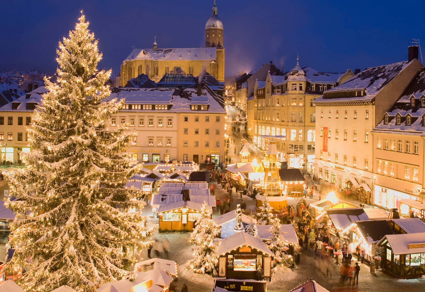 German 桌布 壁紙 — 一棵被雪覆蓋的聖誕樹在夜晚的繁忙冬季市場廣場上溫暖地發光，周圍有歷史建築和節日攤位。