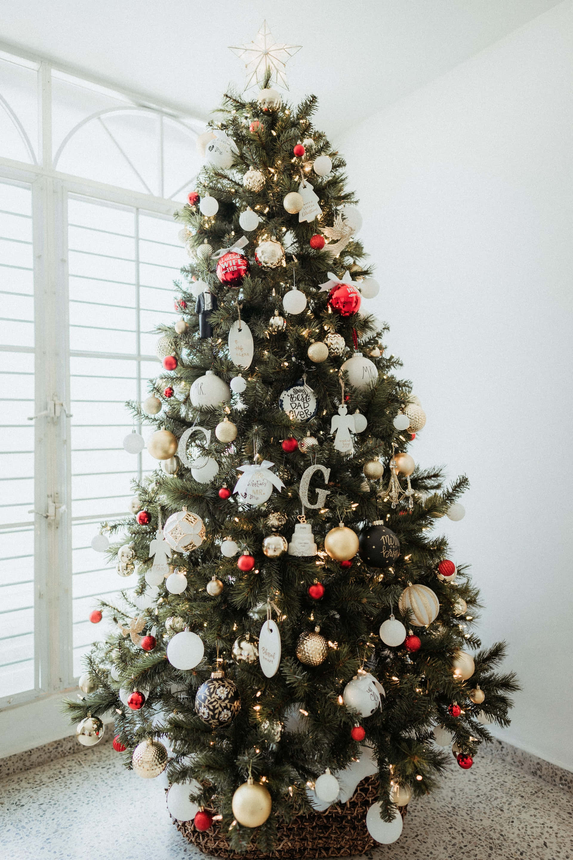 Aesthetic Christmas Tree 桌布 壁紙 — 一棵裝飾精美的聖誕樹，綴以白色、金色和紅色的飾物，優雅地矗立在一個有白色百葉窗的明亮房間中。