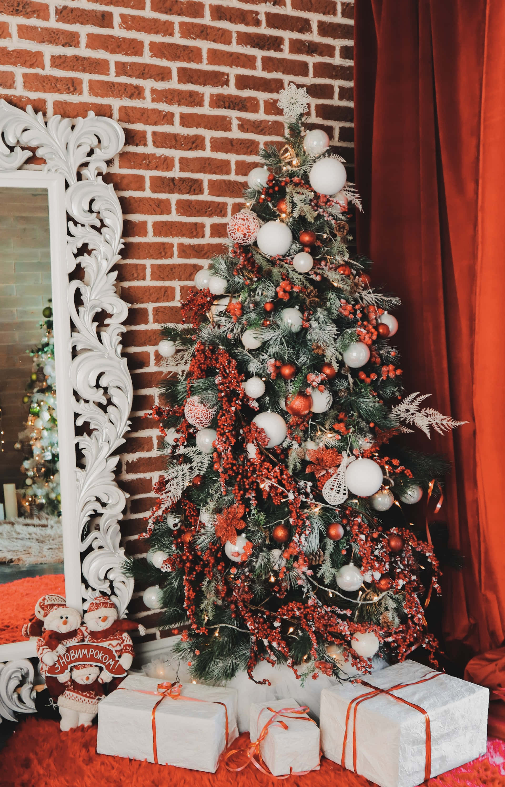 Aesthetic Christmas Tree 桌布 壁紙 — 一棵裝飾華麗的聖誕樹，帶有紅色漿果、白色裝飾品和雪花，矗立在白色華麗鏡子旁邊，背景是磚牆。