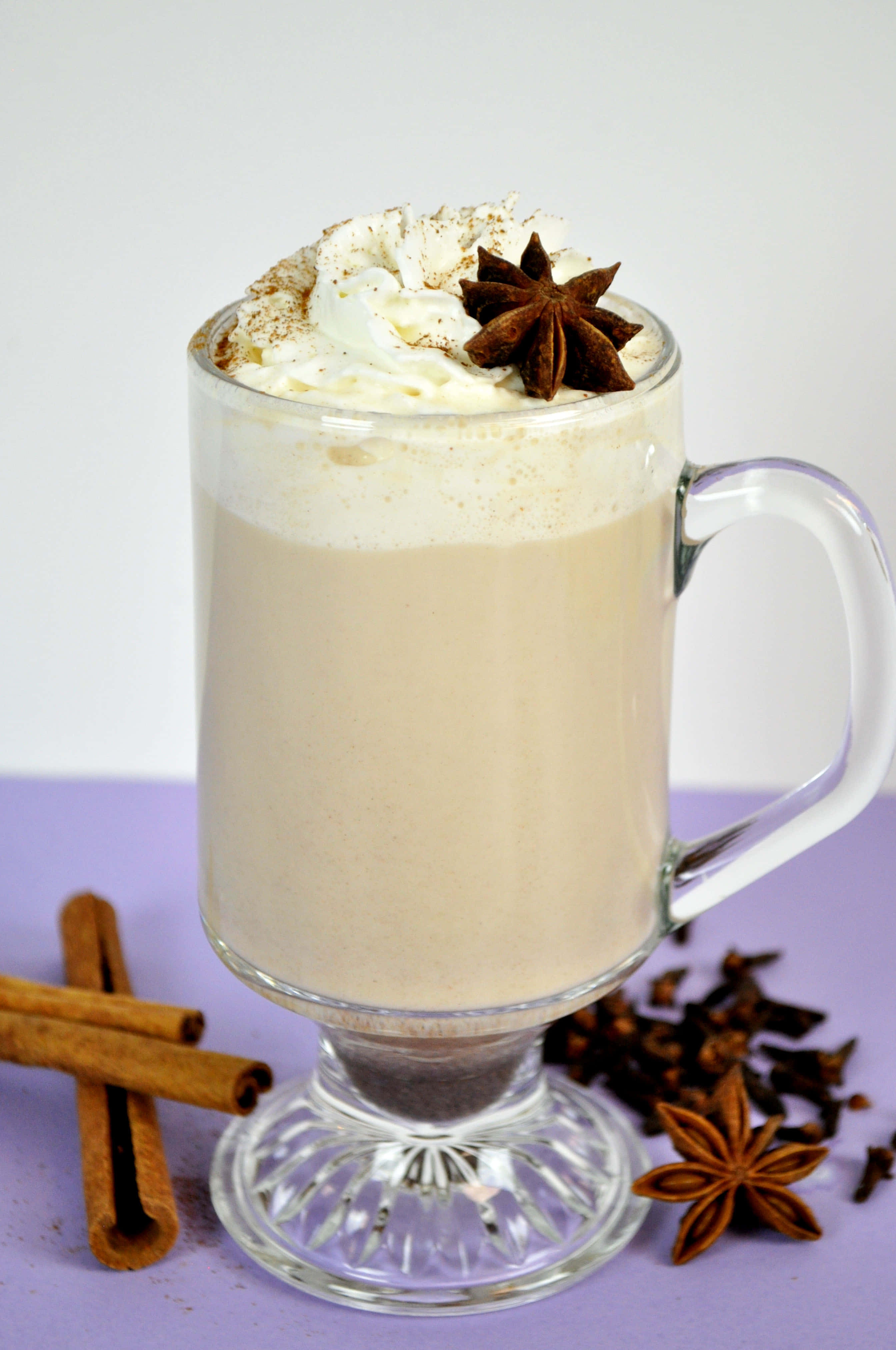 Winter Drinks 桌布 壁紙 — 一款奶油狀的香料飲品，上面放有鮮奶油和八角，放在玻璃杯中，周圍有肉桂棒和丁香，置於紫色表面上。