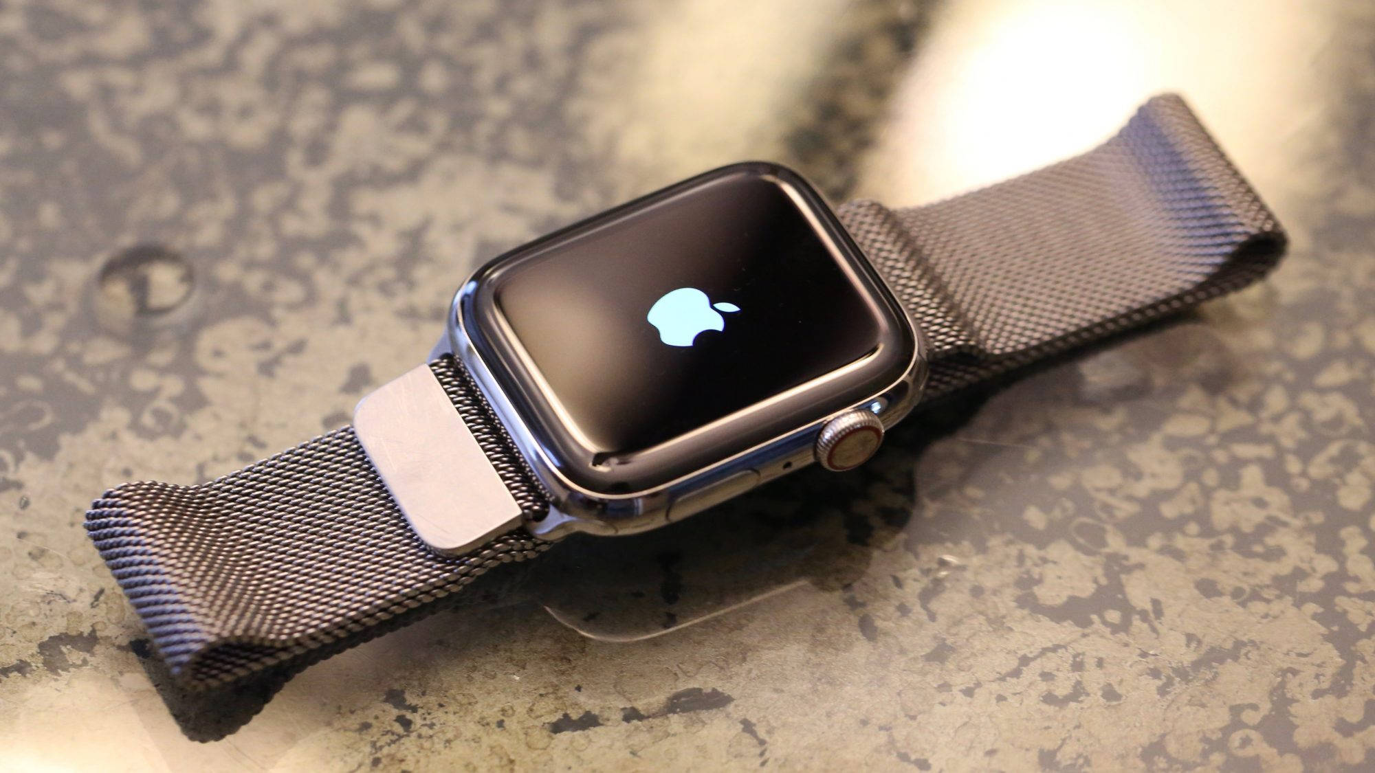 Apple Watch 動態 桌布 壁紙 — 一款時尚的Apple Watch搭配網狀錶帶，放置在斑點檯面上，螢幕閃爍著標誌性標誌。