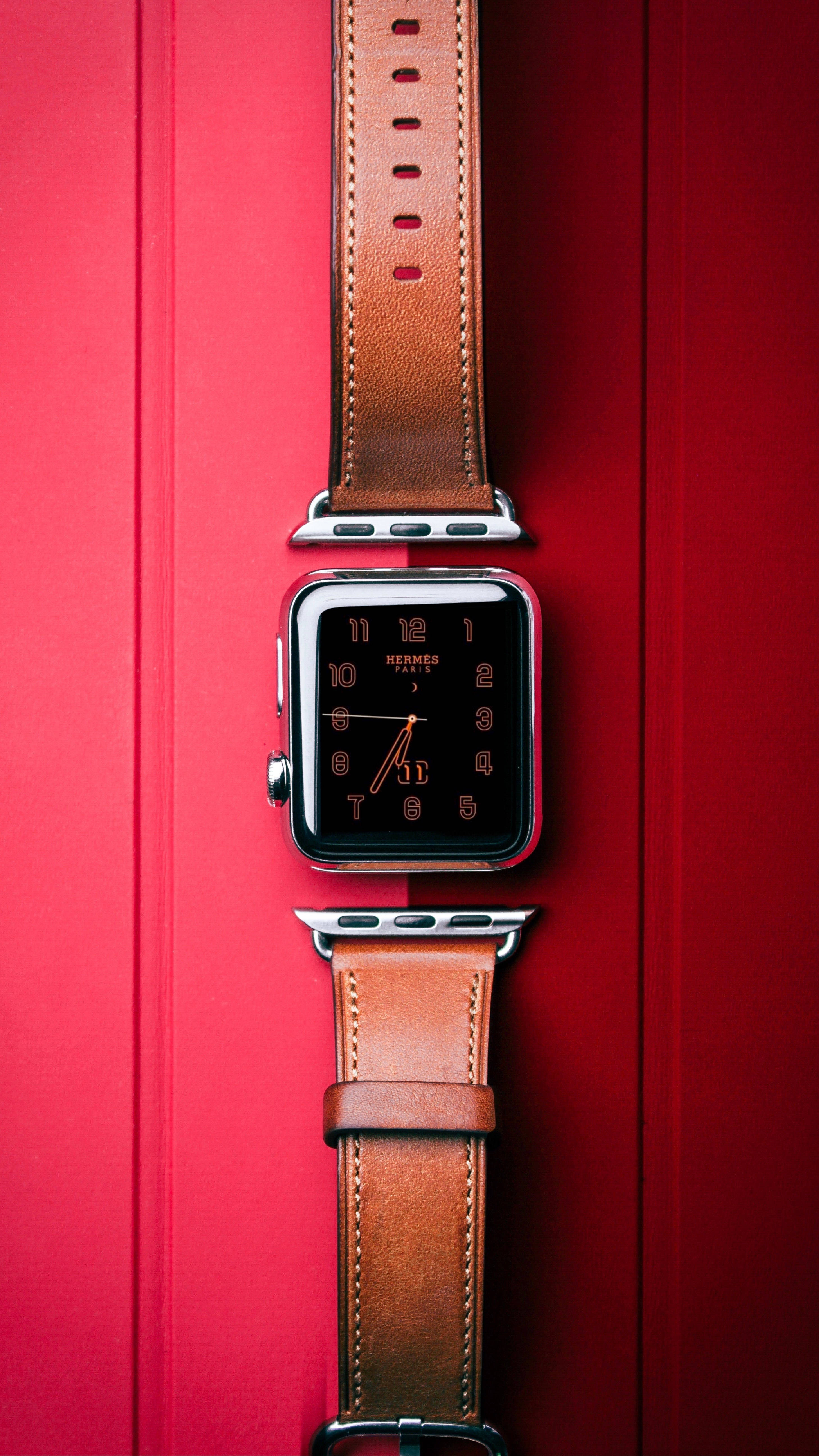 Hermes 桌布 壁紙 — 一款時尚的Apple Watch搭配棕色皮革Hermès錶帶，垂直放置在鮮明的紅色紋理背景上。