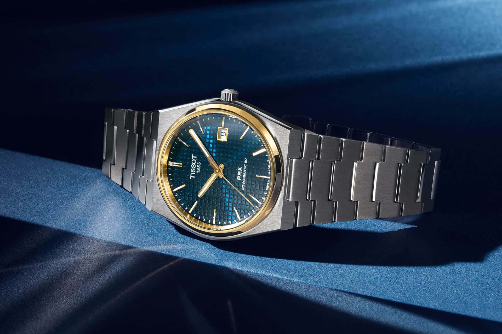 藍色與金色 桌布 壁紙 — 一款時尚的Tissot PRX手錶，藍色錶盤搭配金色點綴，在戲劇性燈光下優雅地放置在深藍色緞面布料上。