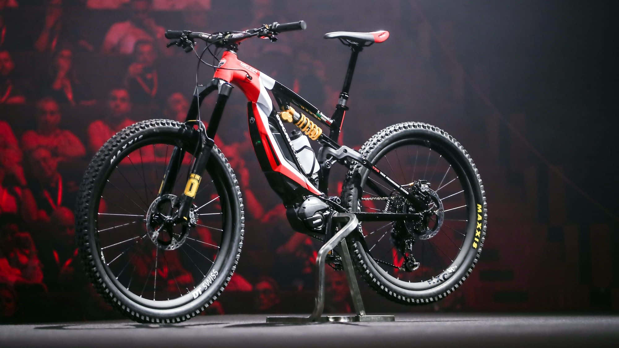 Electric Bike 桌布 壁紙 — 一款時尚的紅黑電動登山車在產品發表會上展示，背後有紅色燈光下的模糊觀眾。