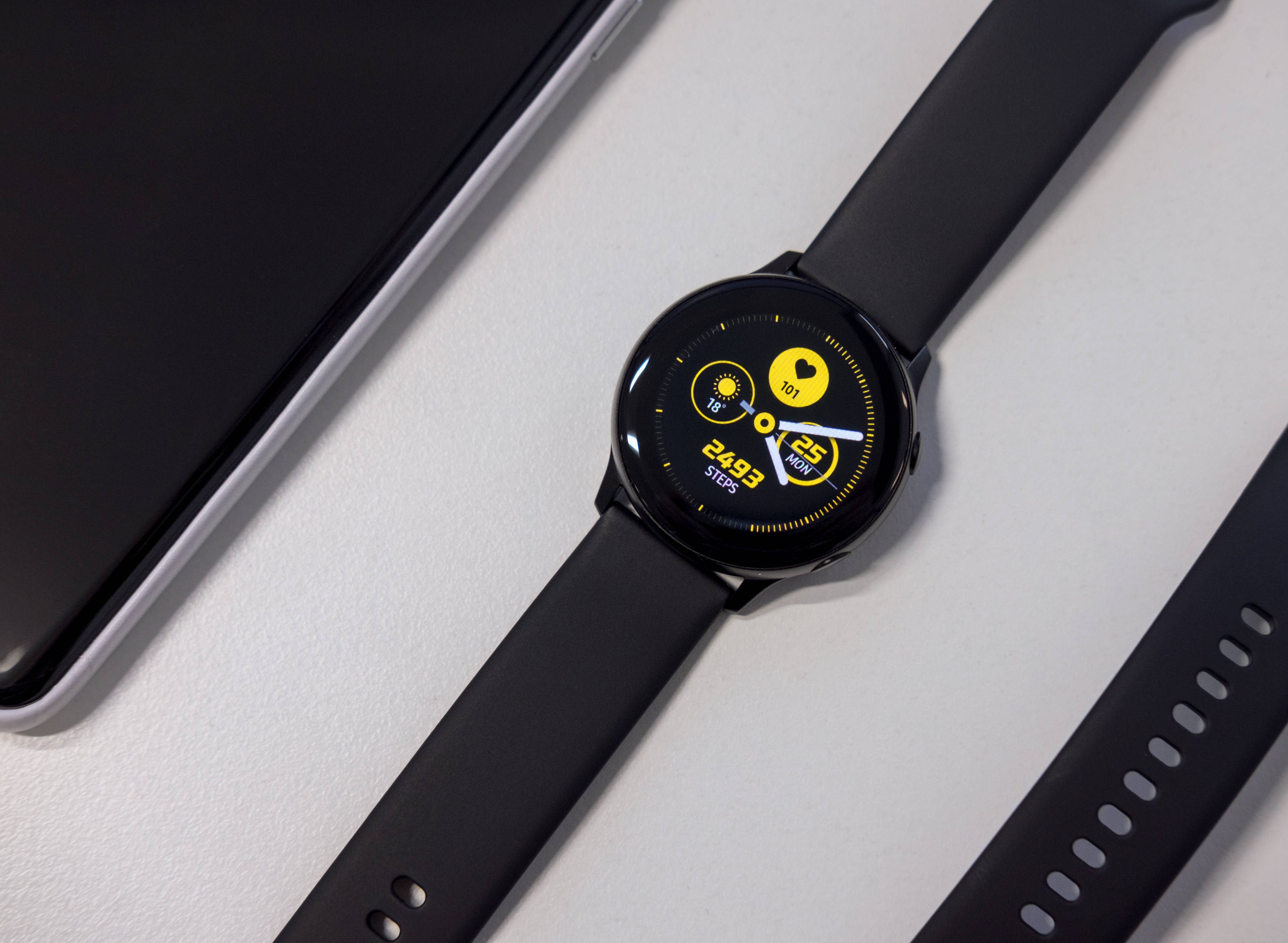 Galaxy S10 桌布 壁紙 — 一款時尚的黑色智慧手錶，帶有鮮豔的黃色健身顯示螢幕，放在極簡白色表面上的智慧型手機旁邊。