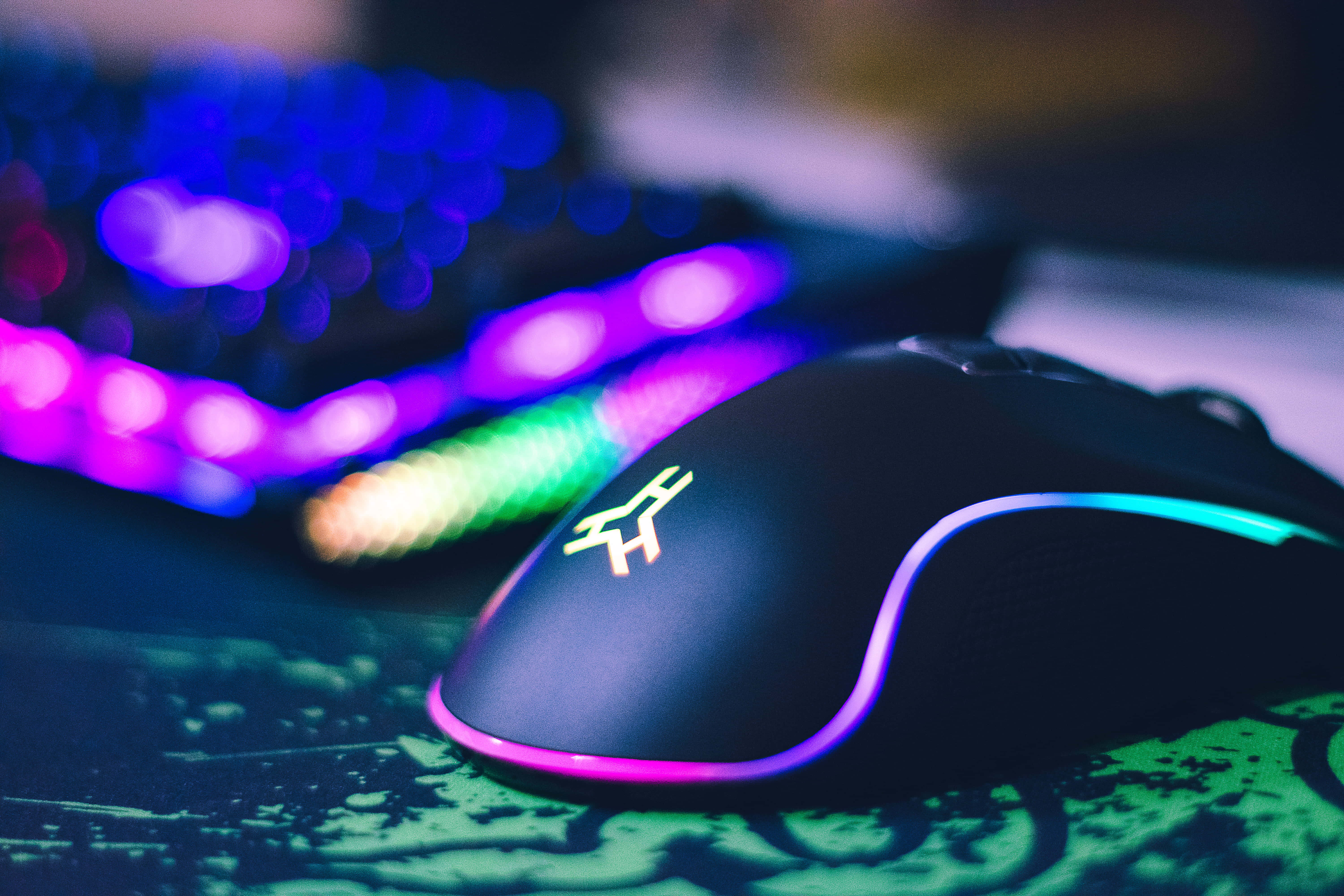 Gaming Mouse 桌布 壁紙 — 一款時尚黑色電競滑鼠帶有發光RGB燈效，放置在圖案滑鼠墊上，背景有模糊鍵盤。