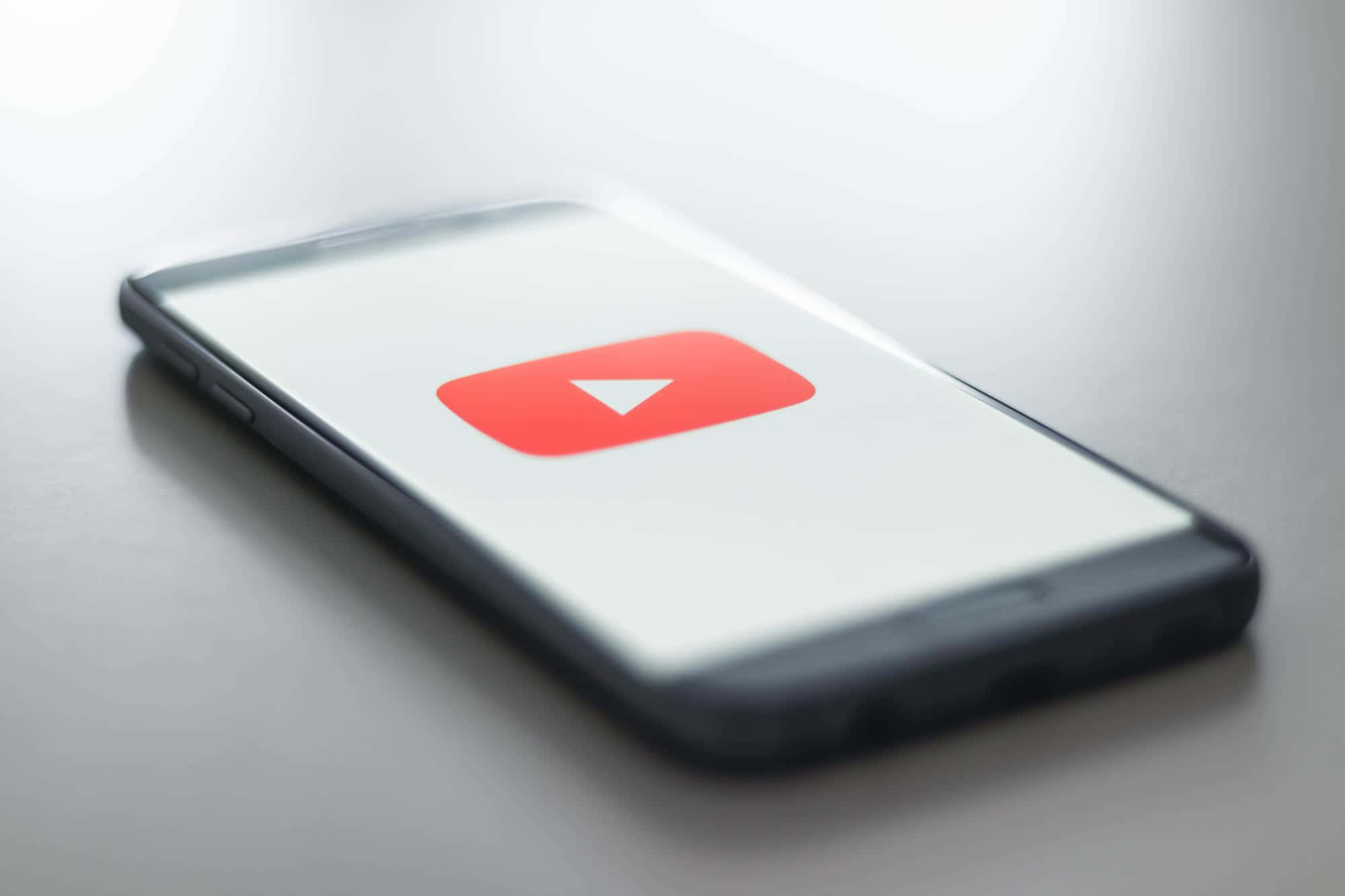 YouTube 桌布 壁紙 — 一款纖薄的智慧型手機在柔和的灰色背景上，以簡約的白色螢幕顯示標誌性的紅色 YouTube 播放按鈕標誌。