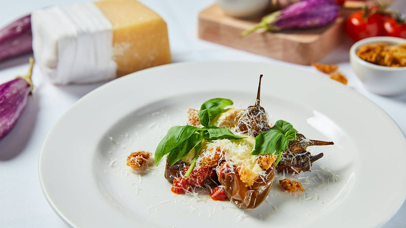 Healthy Food 桌布 壁紙 — 一款美食菜餚，烤茄子配上羅勒起司和香脆麵包丁，放在白色盤子上，背景有起司和番茄。