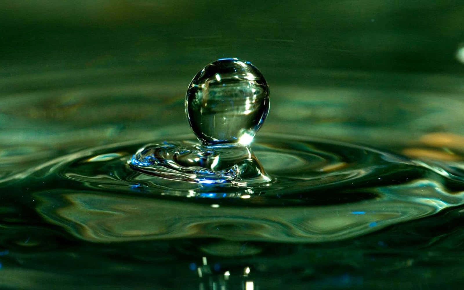Water Drop 桌布 壁紙 — 一滴完美如玻璃的水滴懸浮在漣漪的深綠色水面上，反射著光線。
