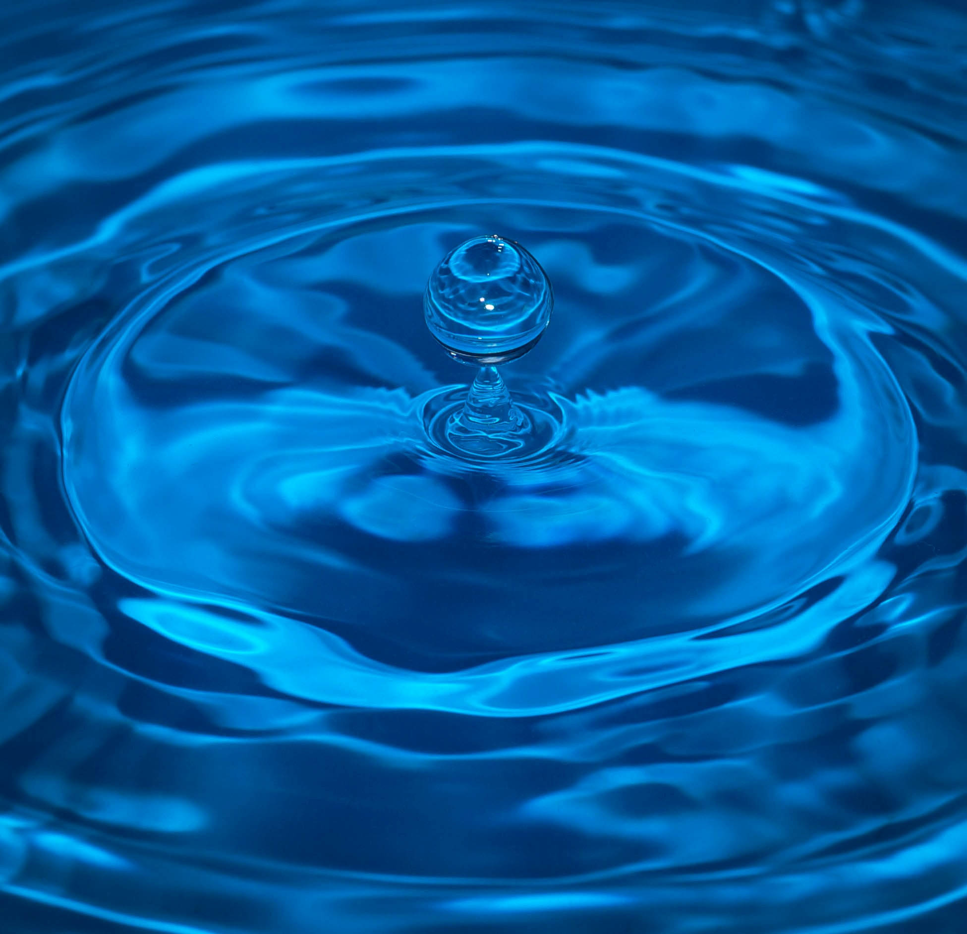 Water Drops 桌布 壁紙 — 一滴完美的藍色水珠懸浮在漣漪池面上方，以慢動作形成同心圓。