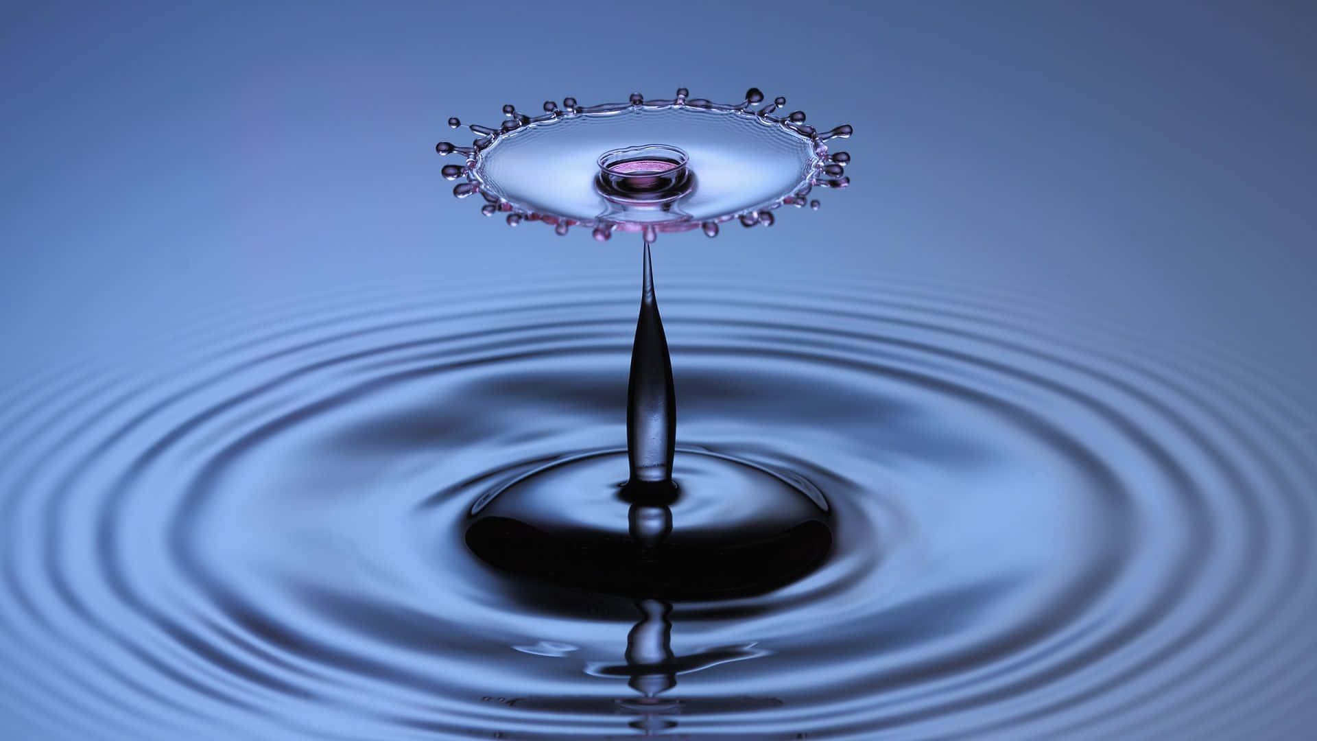 水 桌布 壁紙 — 一滴水懸浮在半空中，在漣漪池面上形成皇冠般的飛濺，背景為冷藍色漸變。