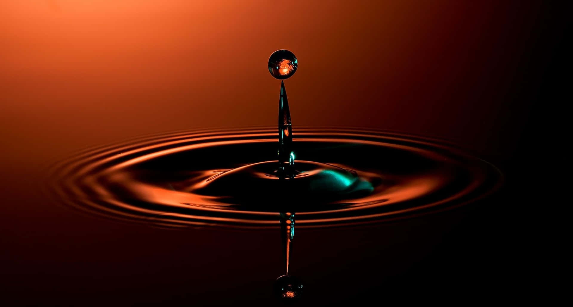 Water Drop 桌布 壁紙 — 一滴水懸浮在黑暗表面上，在溫暖的橙色燈光下形成完美的同心漣漪。