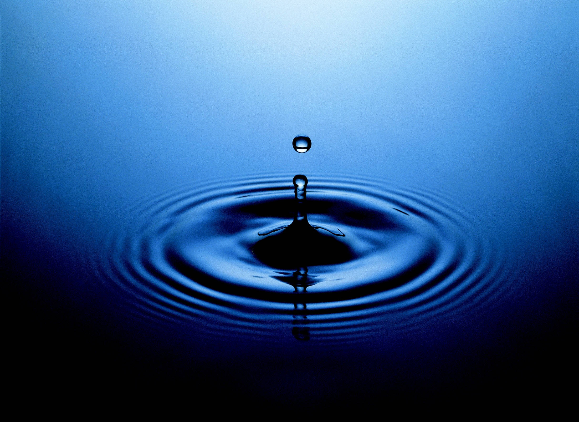 Water Drop 桌布 壁紙 — 一滴水珠懸浮在泛起漣漪的深藍色水池上方，柔和背光照射。