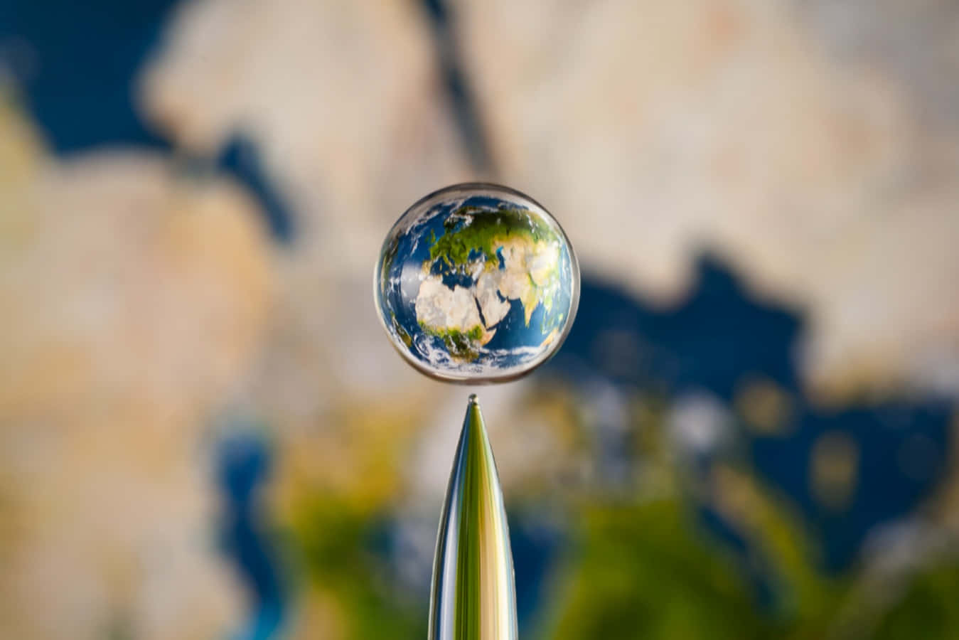 Water Drop 桌布 壁紙 — 一滴水珠棲息在針尖上，完美地反射出整個地球，背景是模糊的地圖。