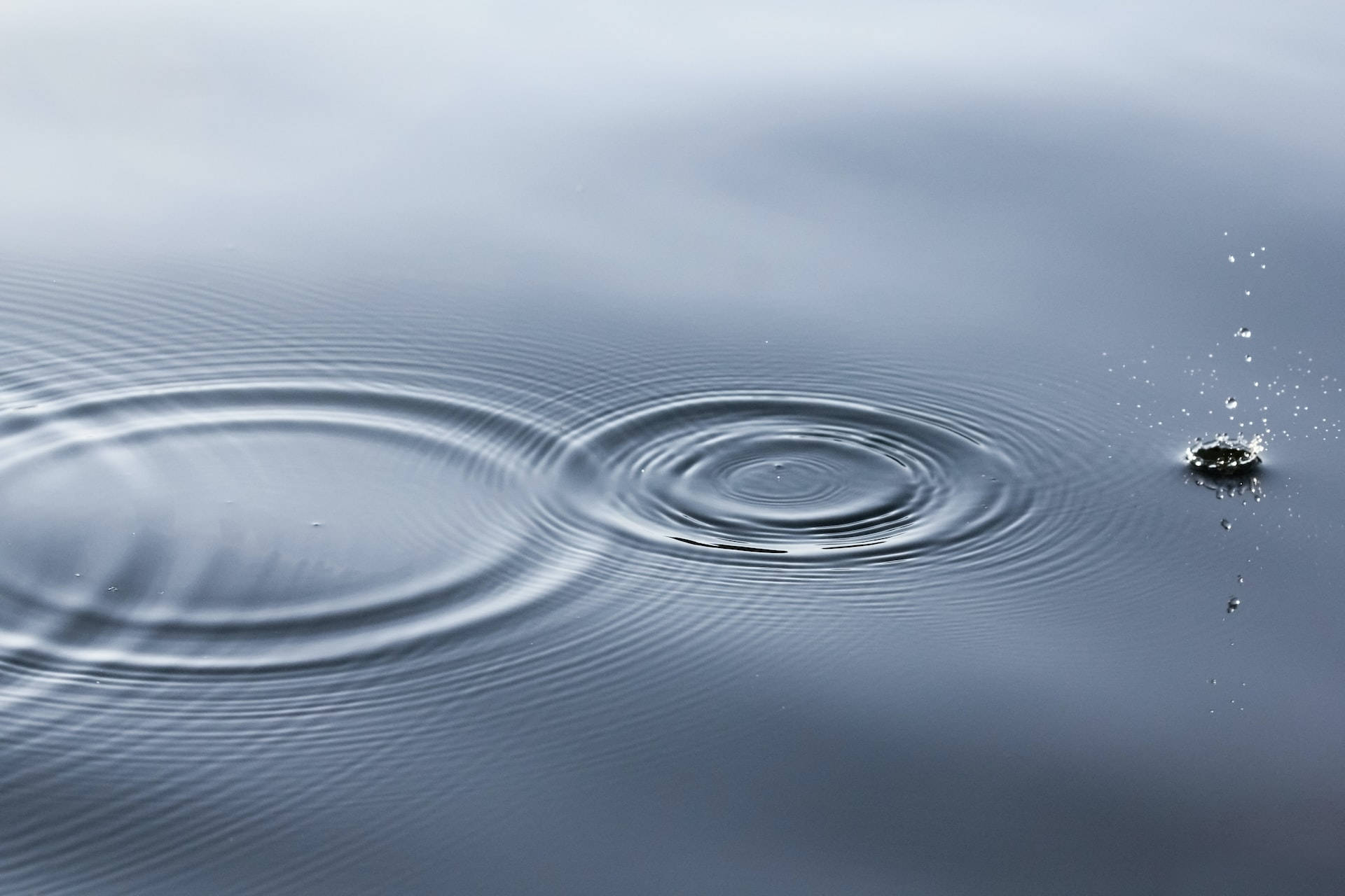 ripple 桌布 壁紙 — 一滴雨滴在平靜的灰色水面上形成擴展的同心漣漪。
