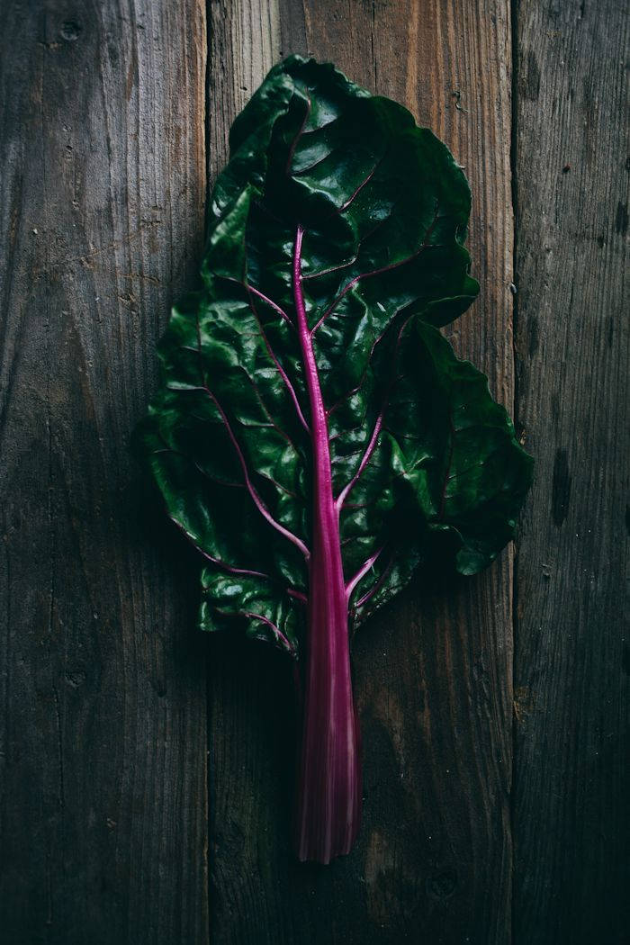 Food Photography 桌布 壁紙 — 一片充滿活力的瑞士甜菜葉，帶有深綠色葉片和鮮豔的洋紅色莖，放在風化的深色木板上。