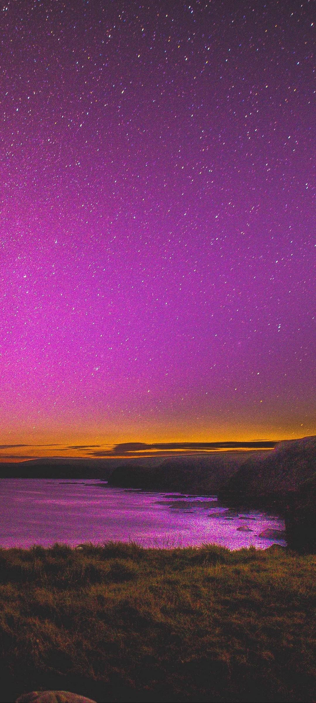 Starry Sky 桌布 壁紙 — 一片充滿活力的紫色星空在暮色中照耀著平靜的海面，襯托出輪廓分明的山丘和長滿青草的前景。