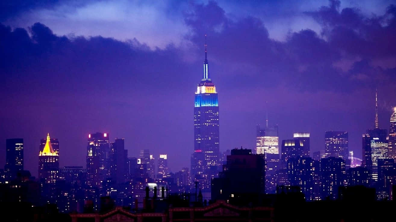1366x768 Empire State Building 桌布 壁紙 — 一片充滿活力的紫色暮色籠罩著紐約市燈火輝煌的天際線，帝國大廈閃爍著藍色和金色的光芒。