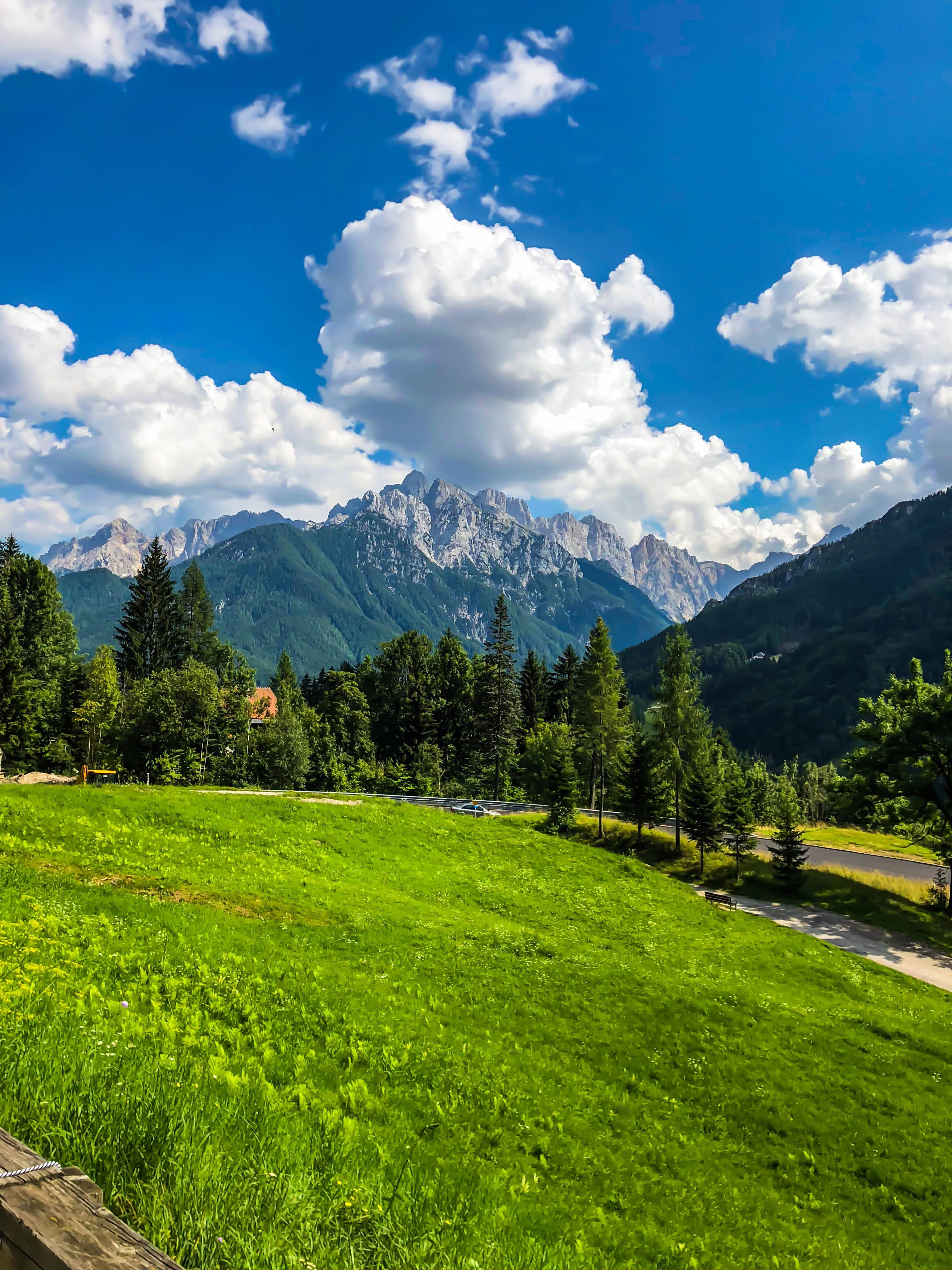 Beautiful Mountain View 桌布 壁紙 — 一片充滿活力的綠色草地，向著一個森林覆蓋的山谷傾斜，山谷中有鋸齒狀的山峰，上方是明亮的藍天，點綴著蓬鬆的白雲。