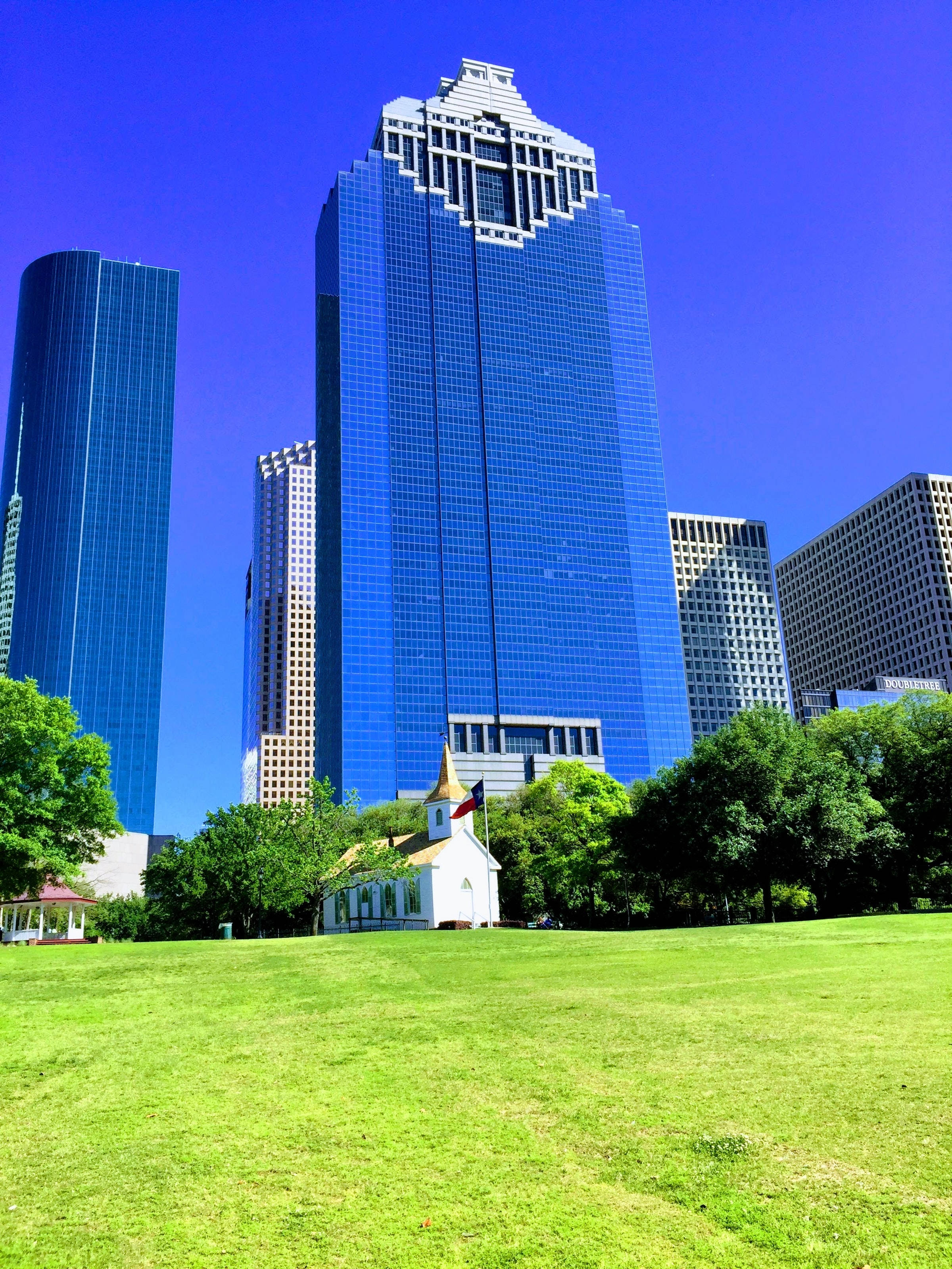 Houston 桌布 壁紙 — 一片充滿活力的綠色草坪延伸在一座歷史悠久的白色教堂前，教堂有金色尖頂，周圍環繞著茂密的樹木和藍色玻璃摩天大樓，在清澈的藍天下。