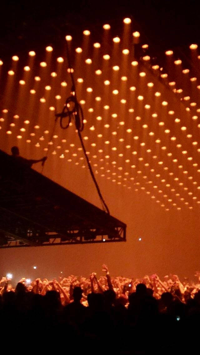 kanye west saint pablo 桌布 壁紙 — 一片剪影般的演唱會觀眾在溫暖橙色舞台燈光的天幕下舉起雙手。