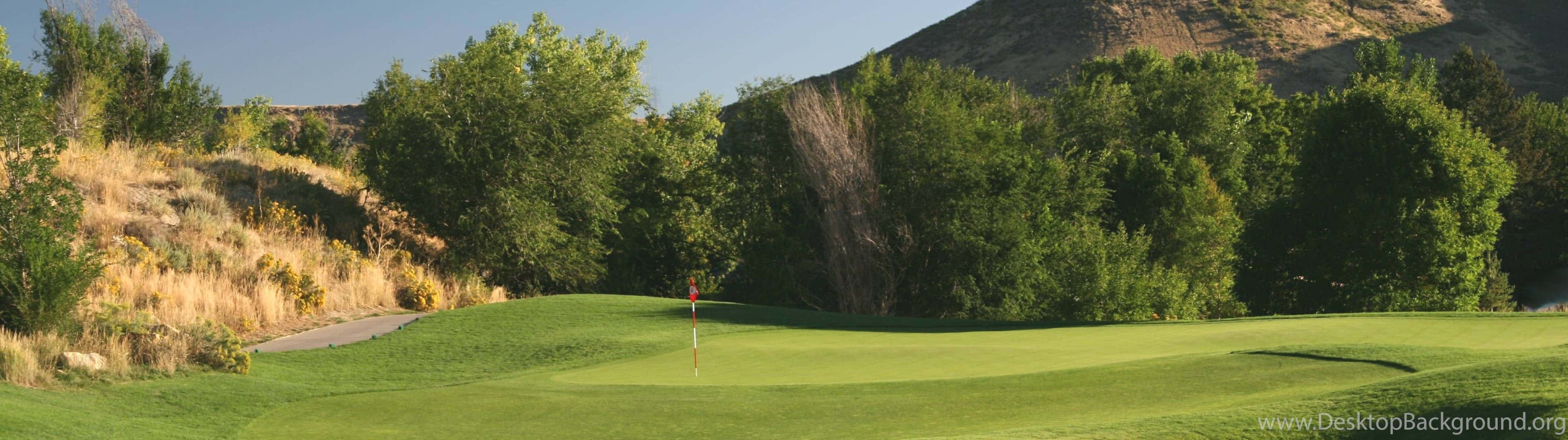 Golf 3840x1080 桌布 壁紙 — 一片寧靜的高爾夫球場果嶺，紅旗點綴其中，四周環繞著茂密的樹木和陽光明媚的山丘，上方是清澈的藍天。