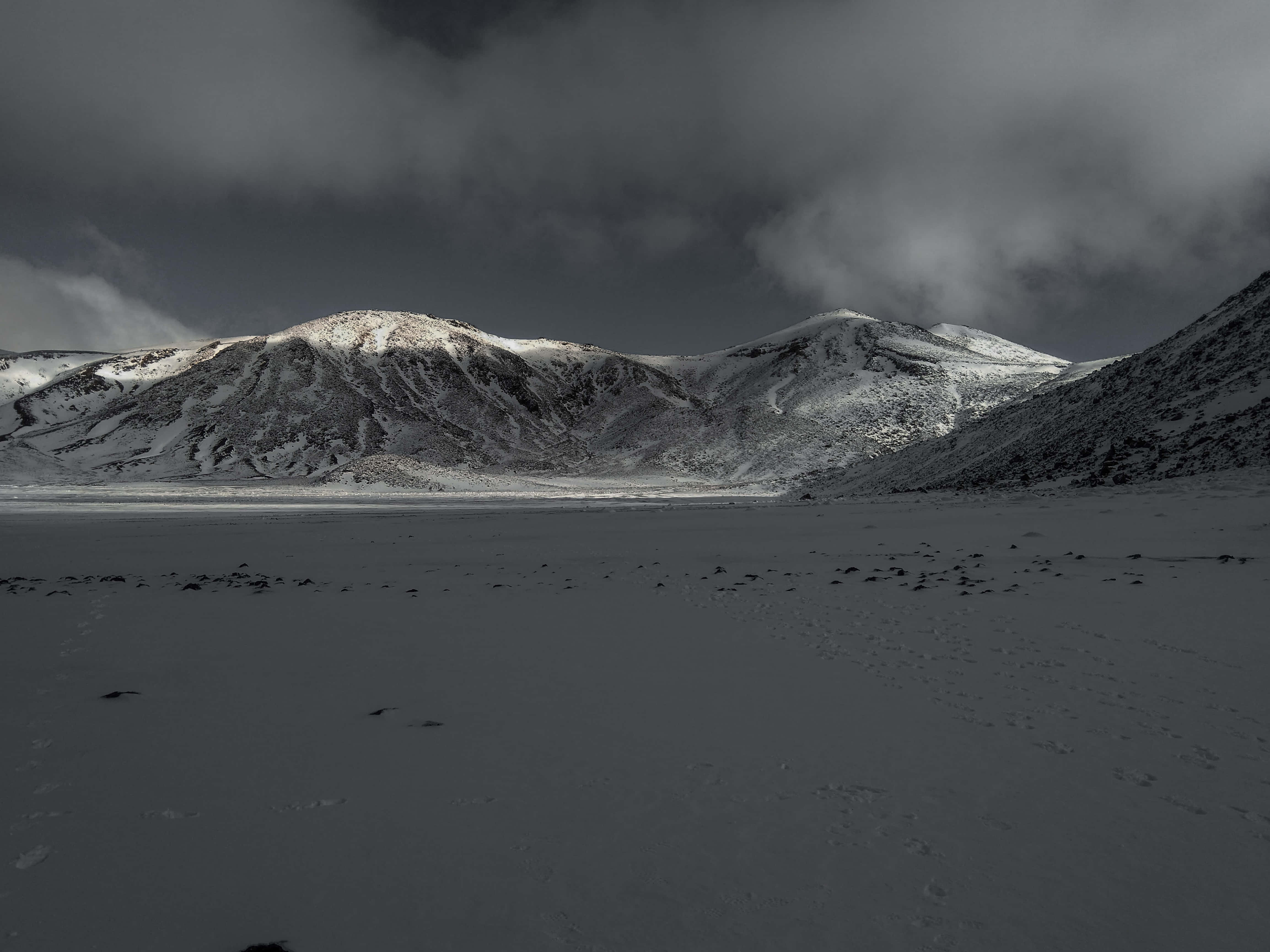 Twitter Header 桌布 壁紙 — 一片廣闊的冰凍湖泊在白雪覆蓋的山脈前延伸，天空陰沉密佈。