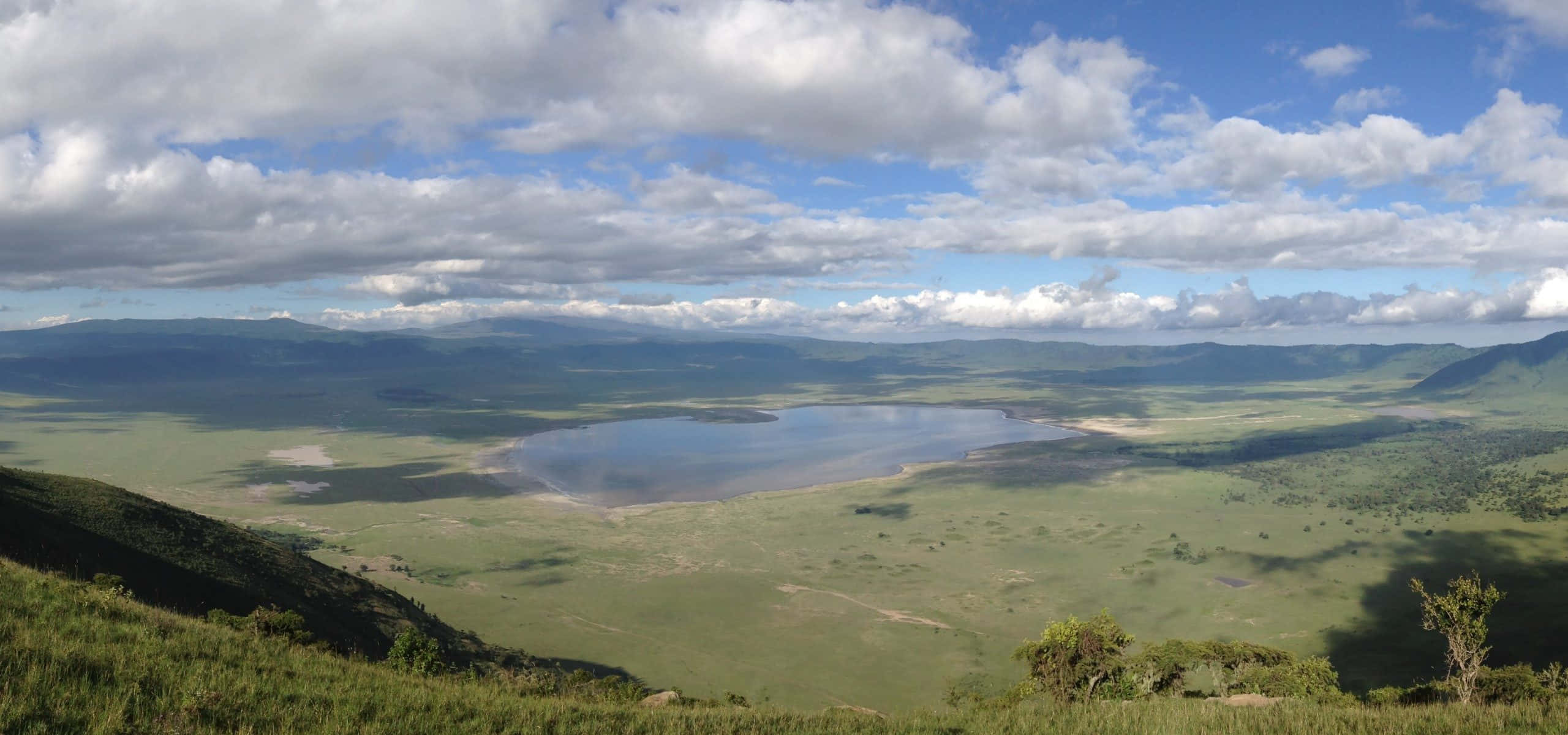 Tanzania 桌布 壁紙 — 一片廣闊的綠色稀樹草原在部分多雲的天空下延伸，山谷中靜臥著一個寧靜的湖泊。