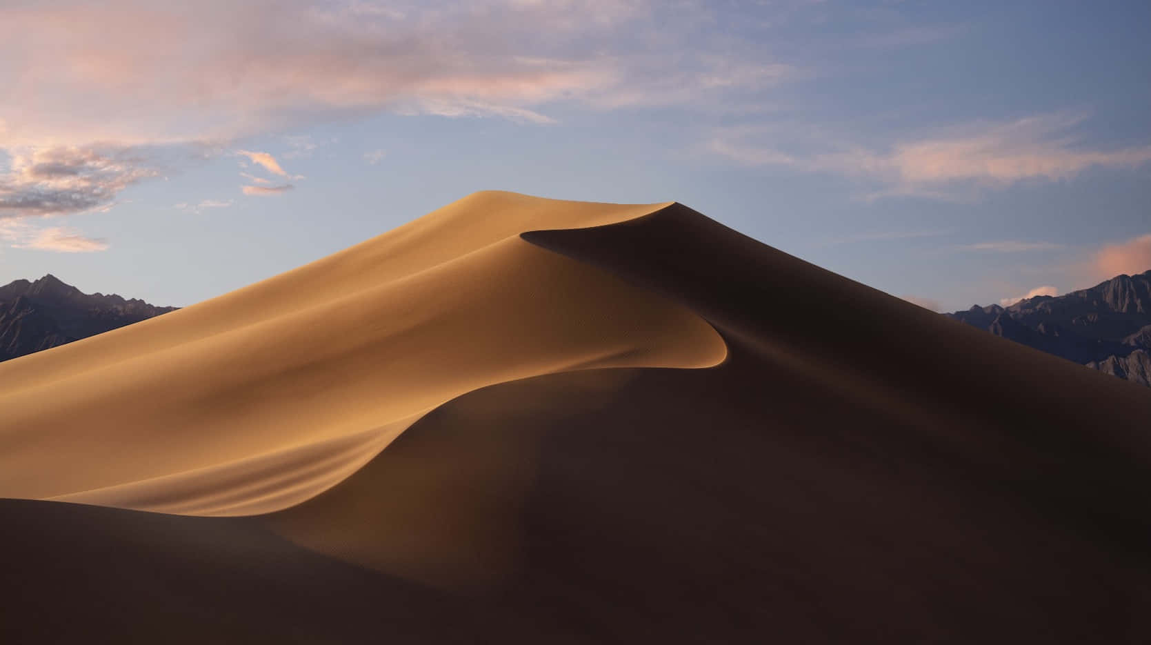 macOS 桌布 壁紙 — 一片廣闊的金色沙丘在柔和的暮色天空下優雅地彎曲，遠山的剪影在背景中浮現。
