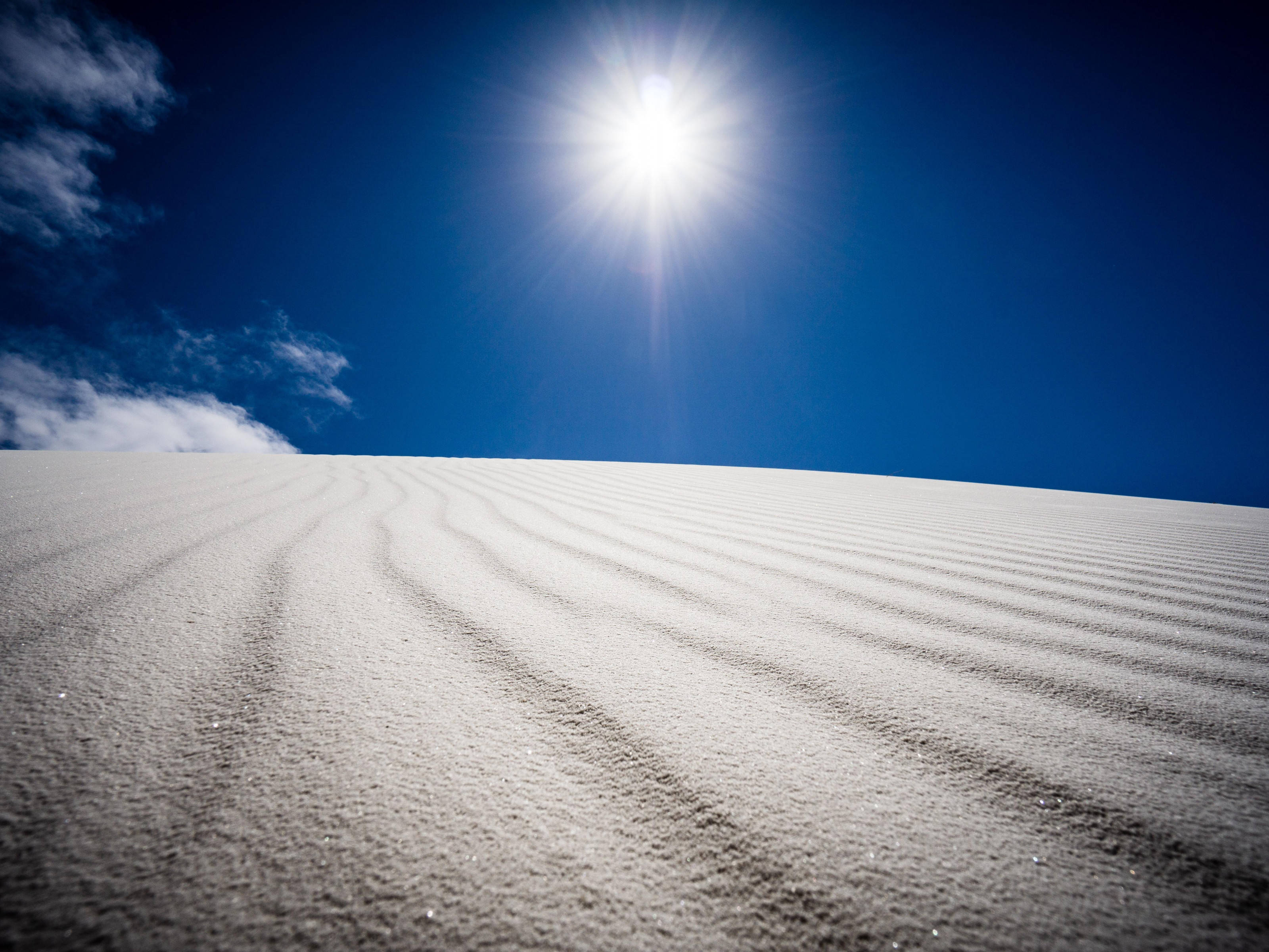 沙漠太陽 桌布 壁紙 — 一片廣闊的雪沙丘在燦爛的陽光下延伸，尖銳的光線穿透深藍色的天空和縷縷雲彩。