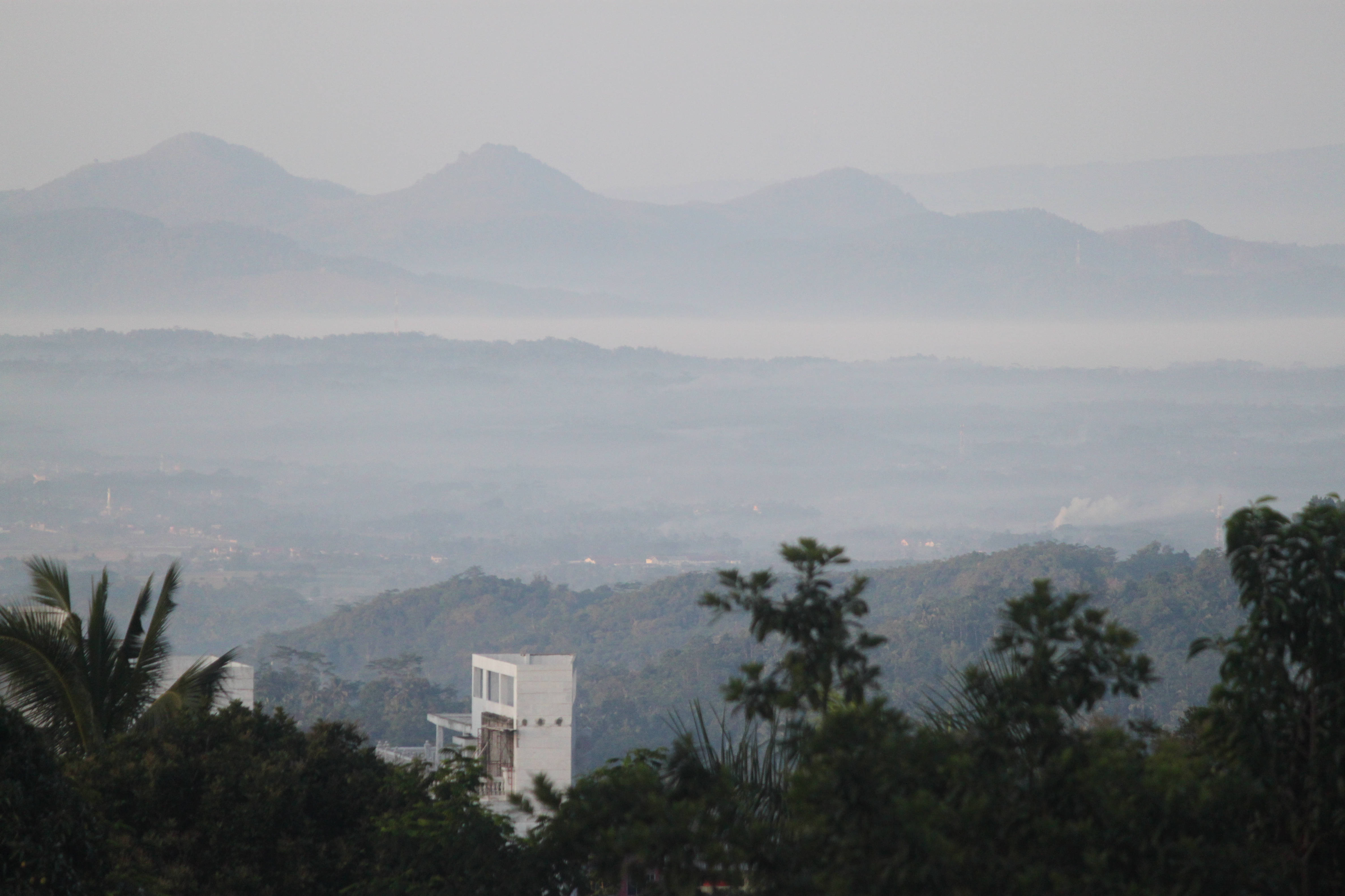 pemandangan 桌布 壁紙 — 一片朦朧的山脈在地平線上延伸，背後是霧氣瀰漫的山谷，前景有零星的建築物和茂密的綠樹。