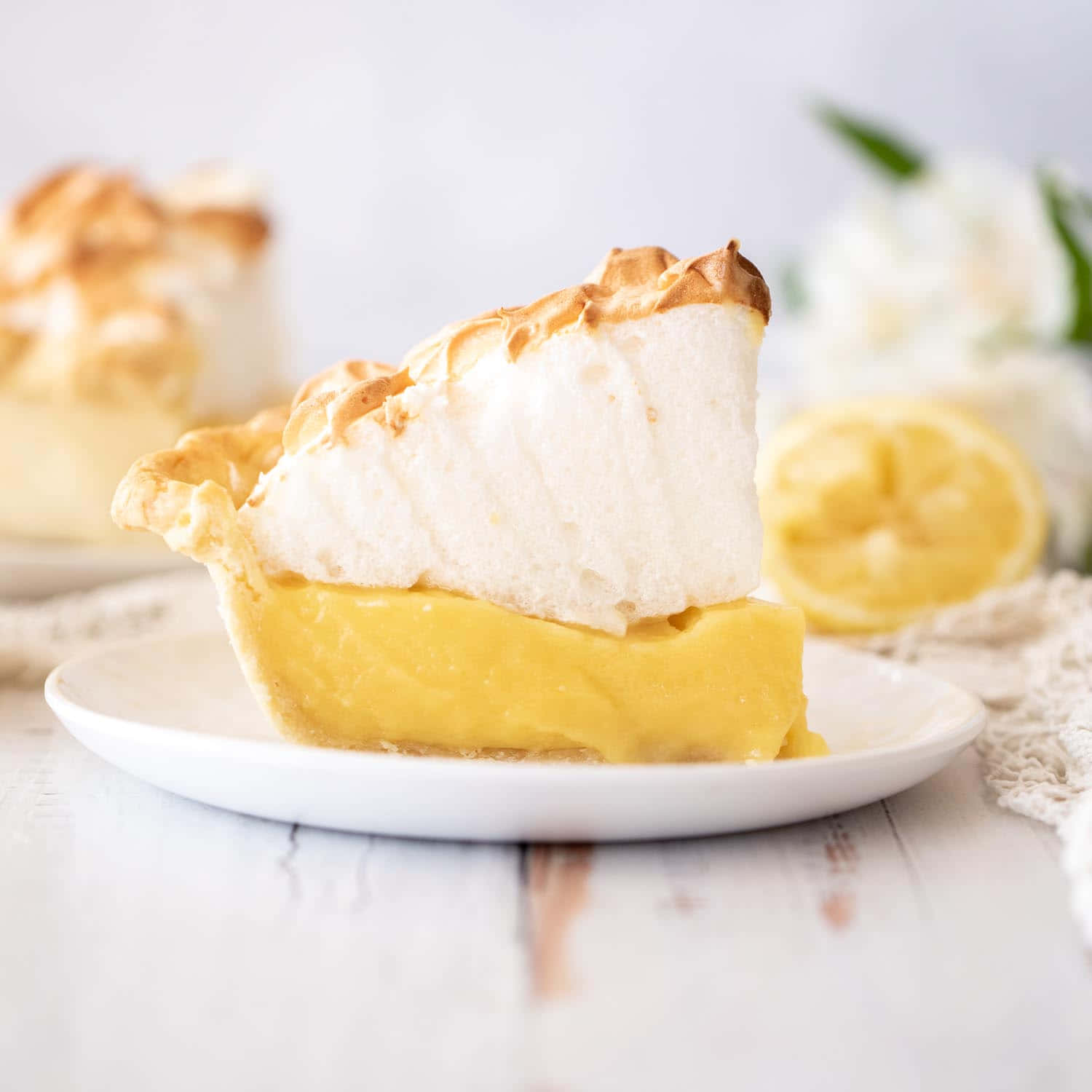 Meringue 桌布 壁紙 — 一片檸檬蛋白派，表面有烤焦的尖峰，放在白色盤子上，旁邊有半個檸檬和白色花朵。