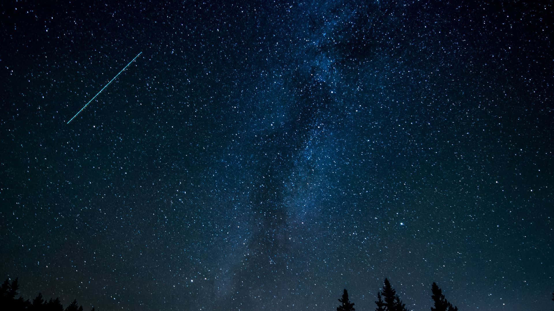 桌布 壁紙 — 一片燦爛的藍色夜空，佈滿星星，銀河在黑暗的松樹剪影上方拱起，一顆流星劃過天際。